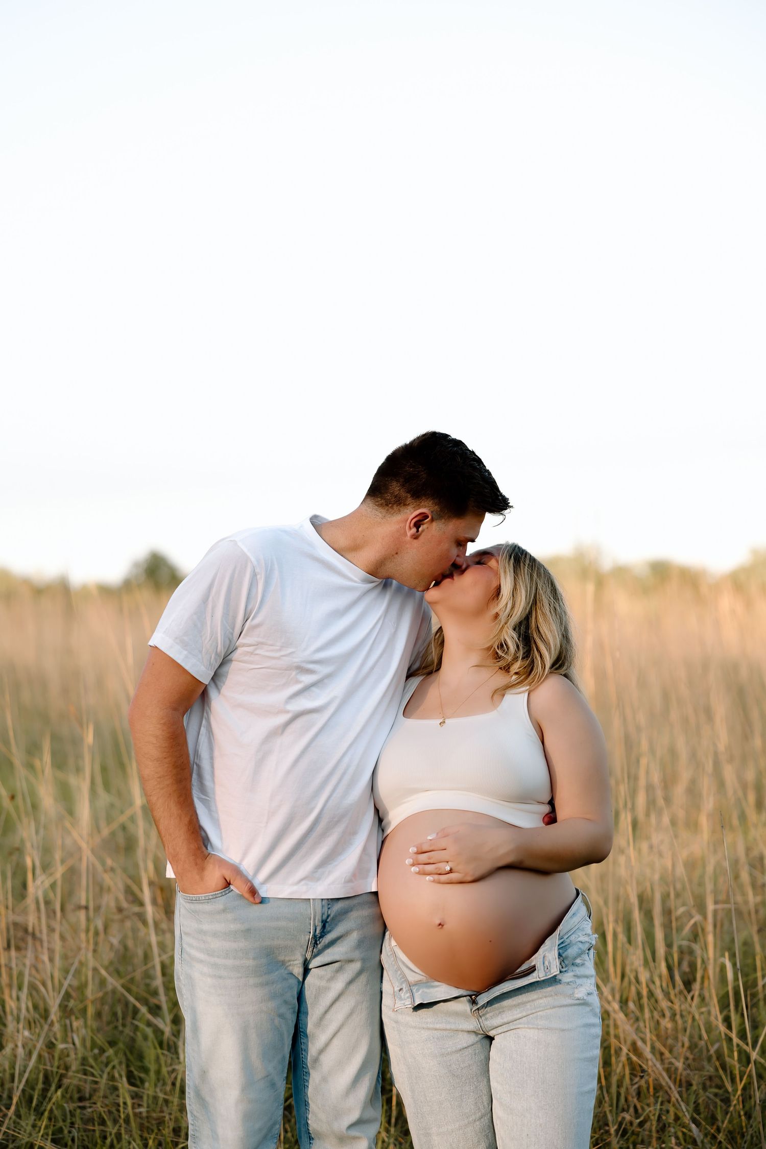 Couple embracing in a sunlit field during maternity photo session showing pregnant belly in white clothing.