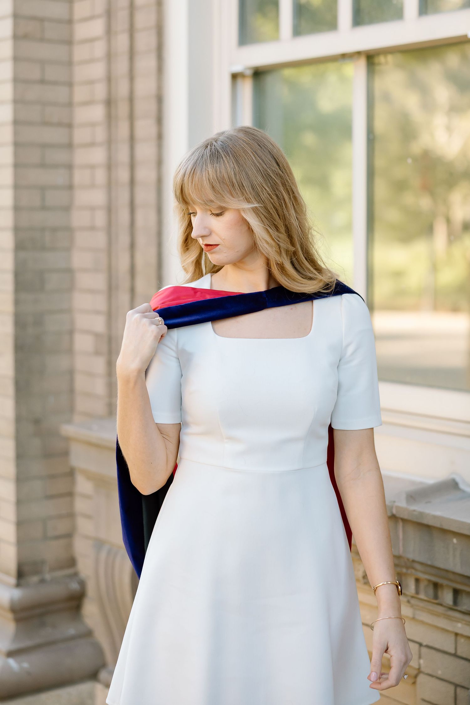 A woman in a white short-sleeved dress drapes a navy blue jacket over her shoulder while standing by a brick building.