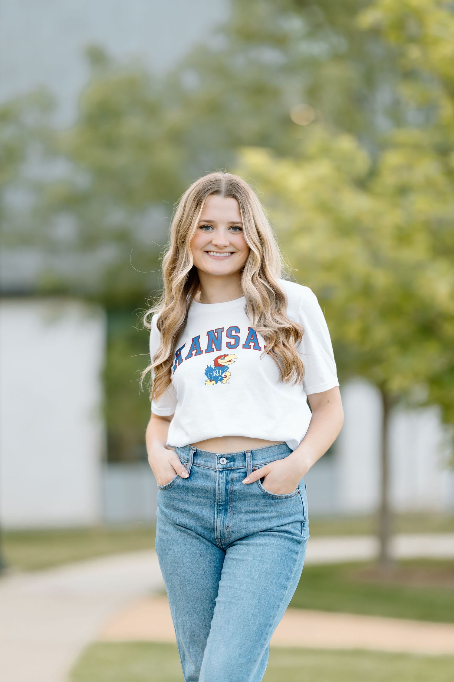 Person in a cropped Kansas t-shirt and blue jeans poses casually outdoors against a blurred natural background.
