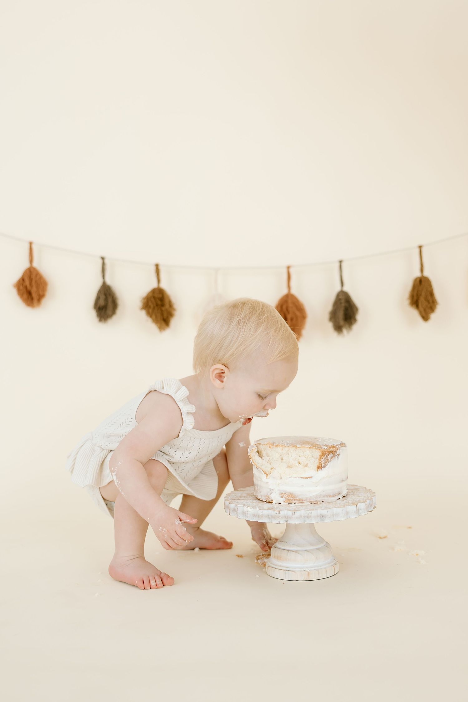 Baby in white romper leans down to investigate cake with dried leaves hanging on wall.