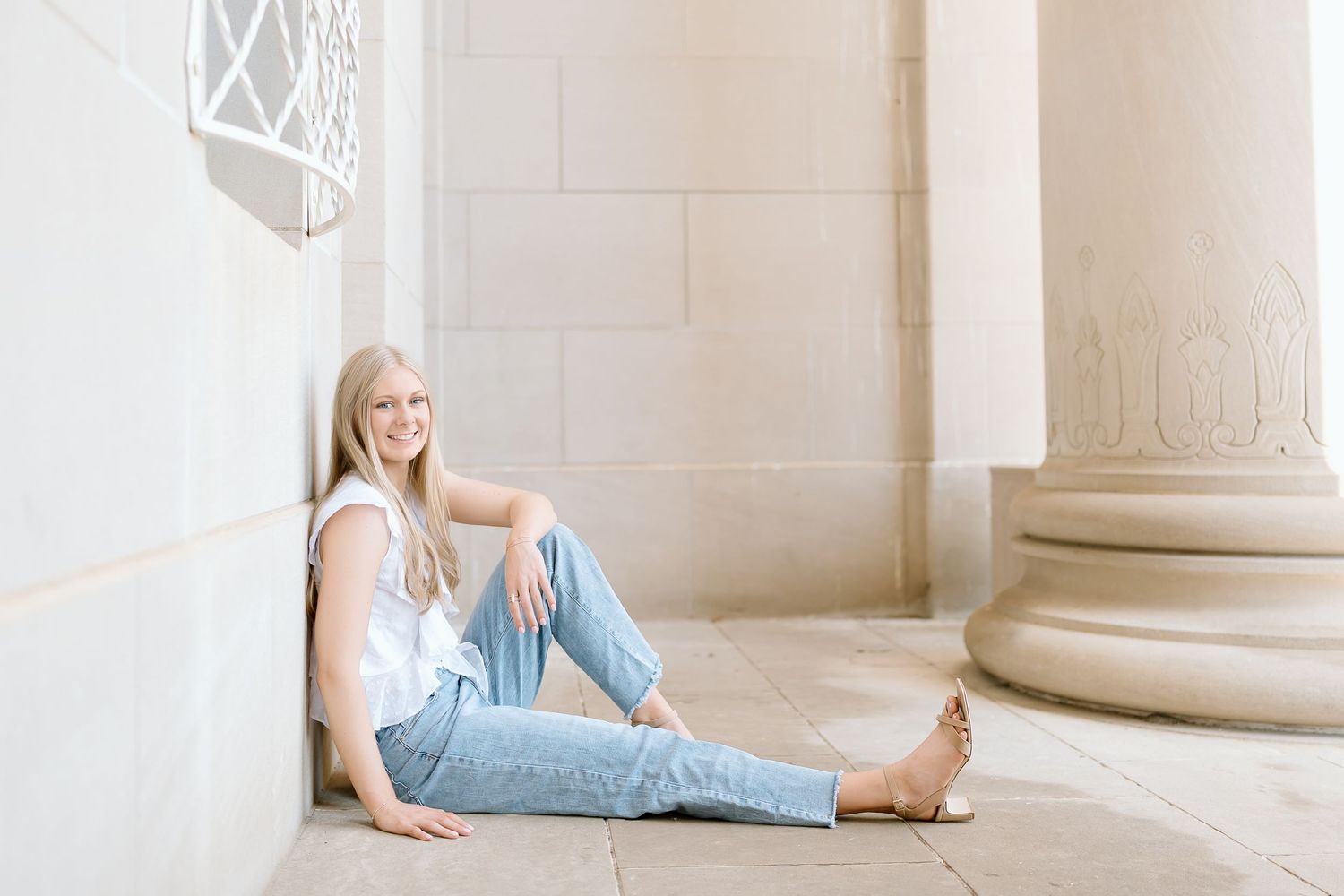A person in light blue jeans and white top sits casually against a stone pillar in an elegant architectural setting.