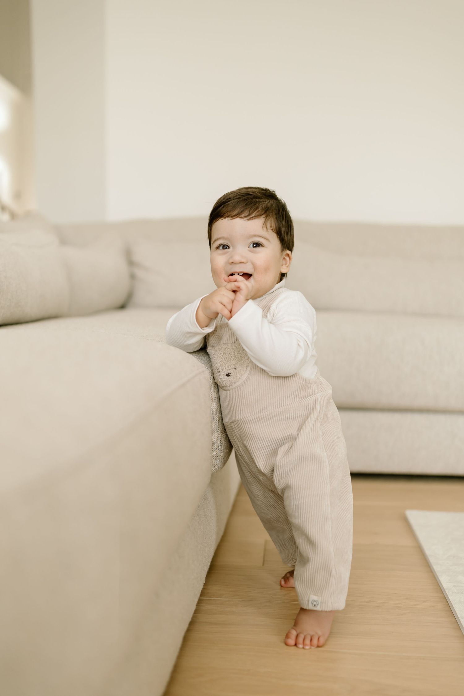A small child in a white outfit crawls and plays on a wooden floor next to a light-colored sofa.