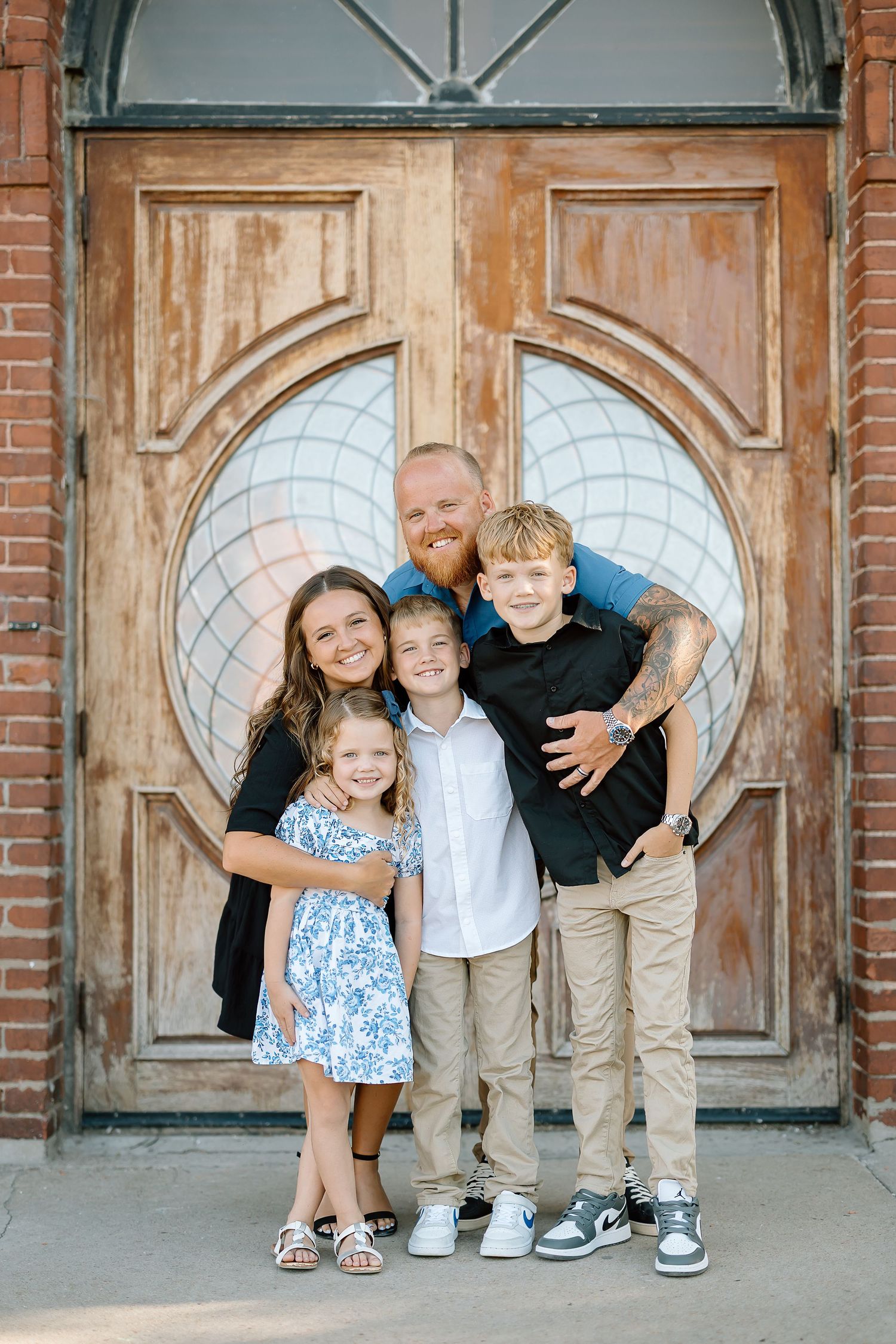 A family poses together in front of a rustic wooden door with a heart-shaped window at a brick church entrance.
