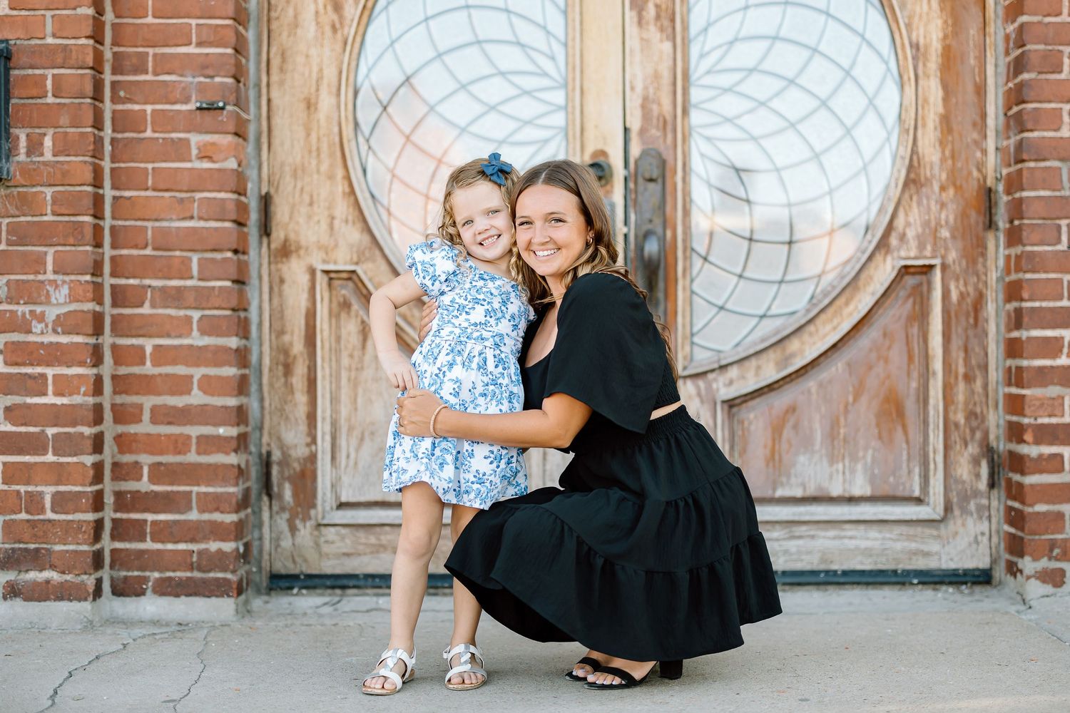 Two people pose in front of a rustic wooden door with circular window designs and brick wall background.