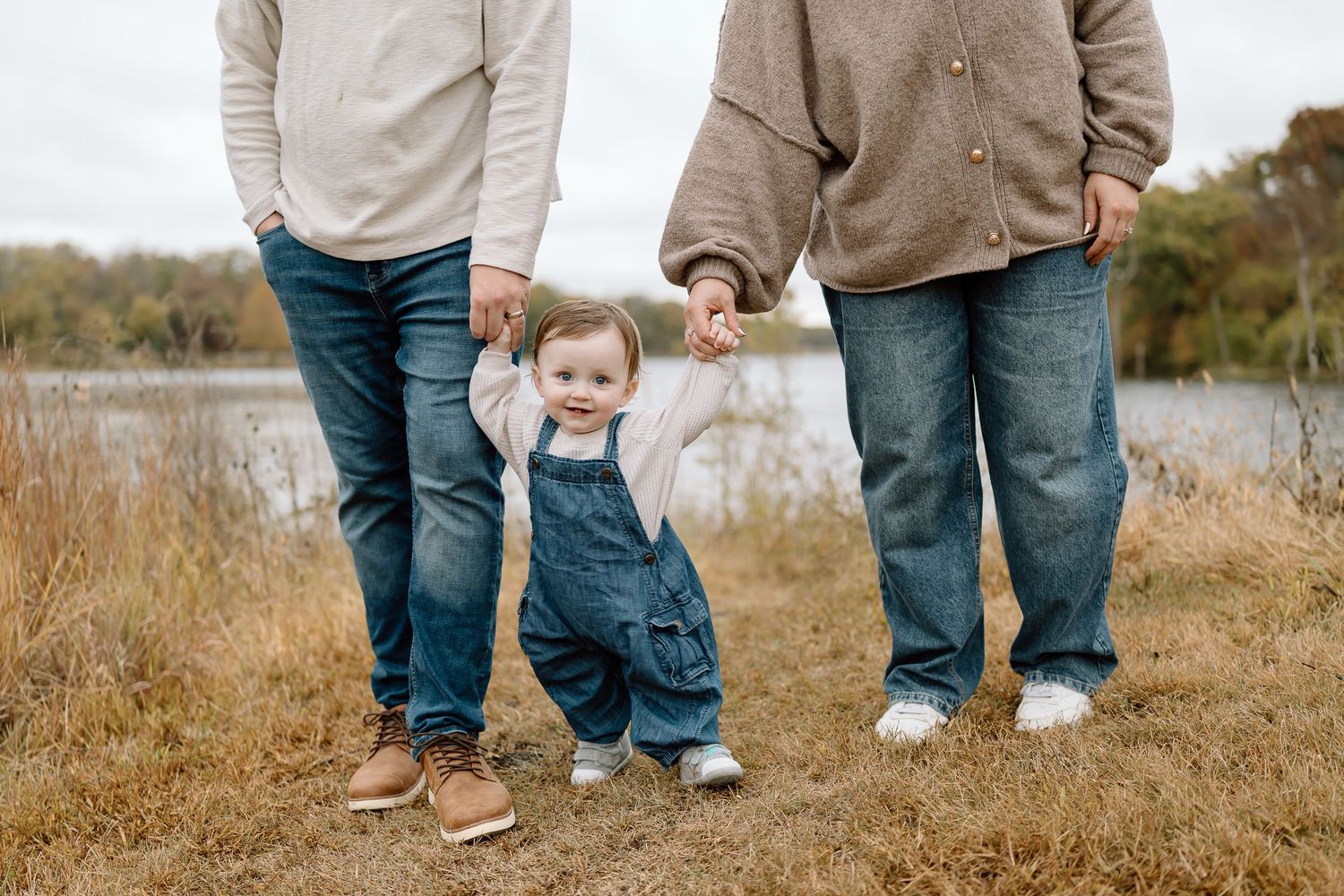 A small child in denim overalls holds hands with adults while standing in tall grass by a lake.