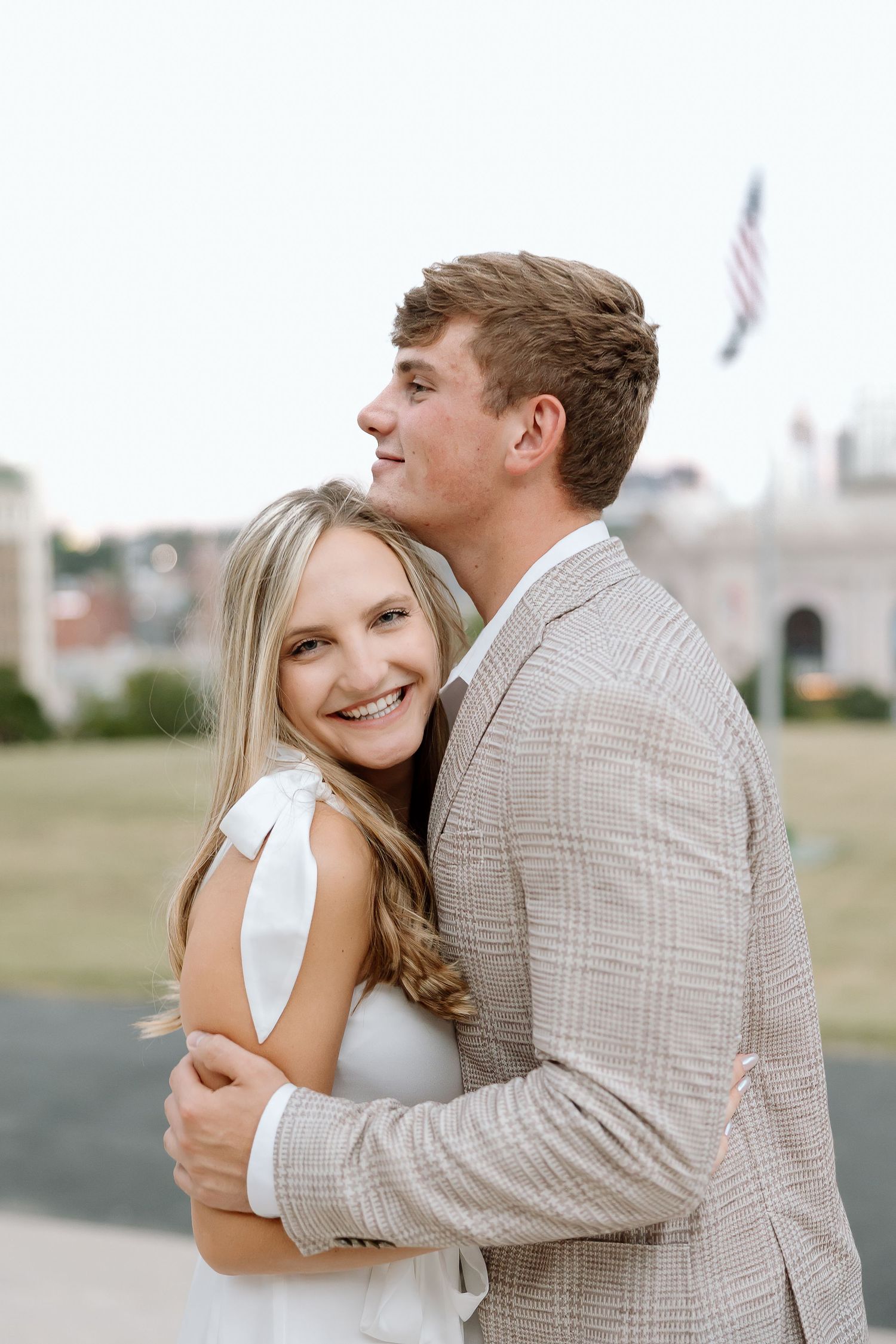 A romantic couple embraces outdoors with an American flag visible in the background.