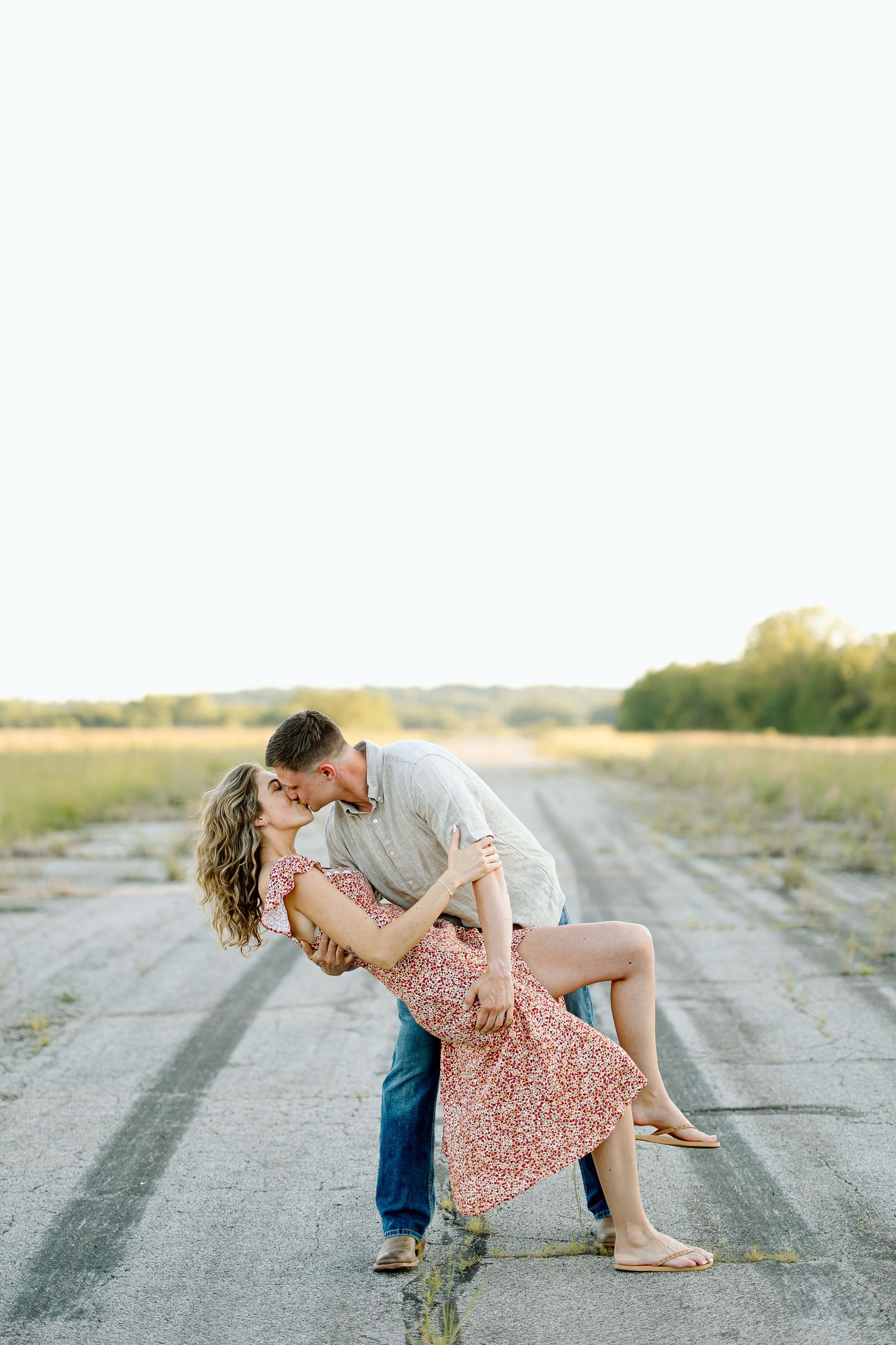 Romantic couple in casual attire share an intimate dance pose on a rural dirt road at sunset.