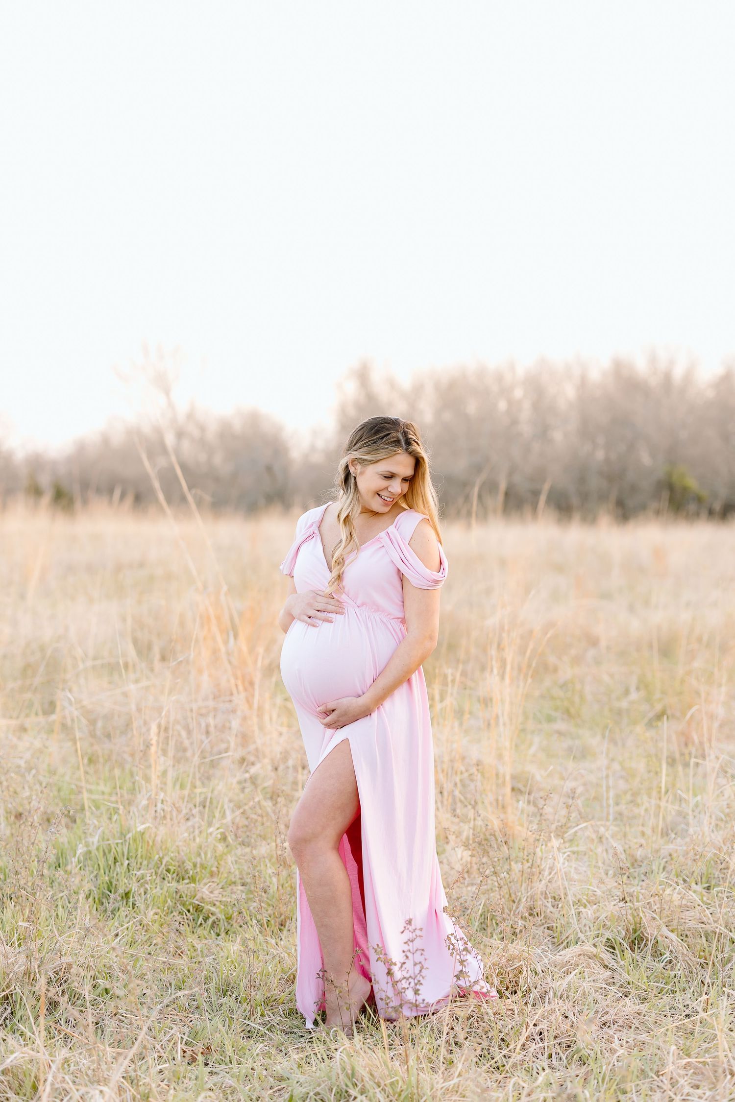 A woman in a flowing pink dress poses gracefully in a sunlit field of tall grass during golden hour.