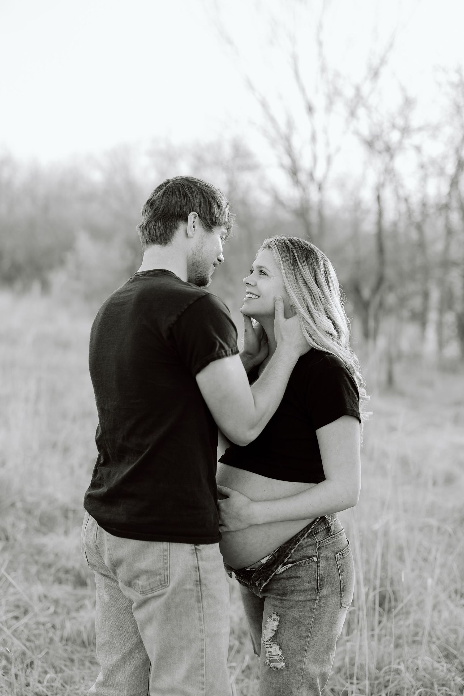 Black and white maternity photo of a couple embracing in a natural outdoor setting with bare trees in background.