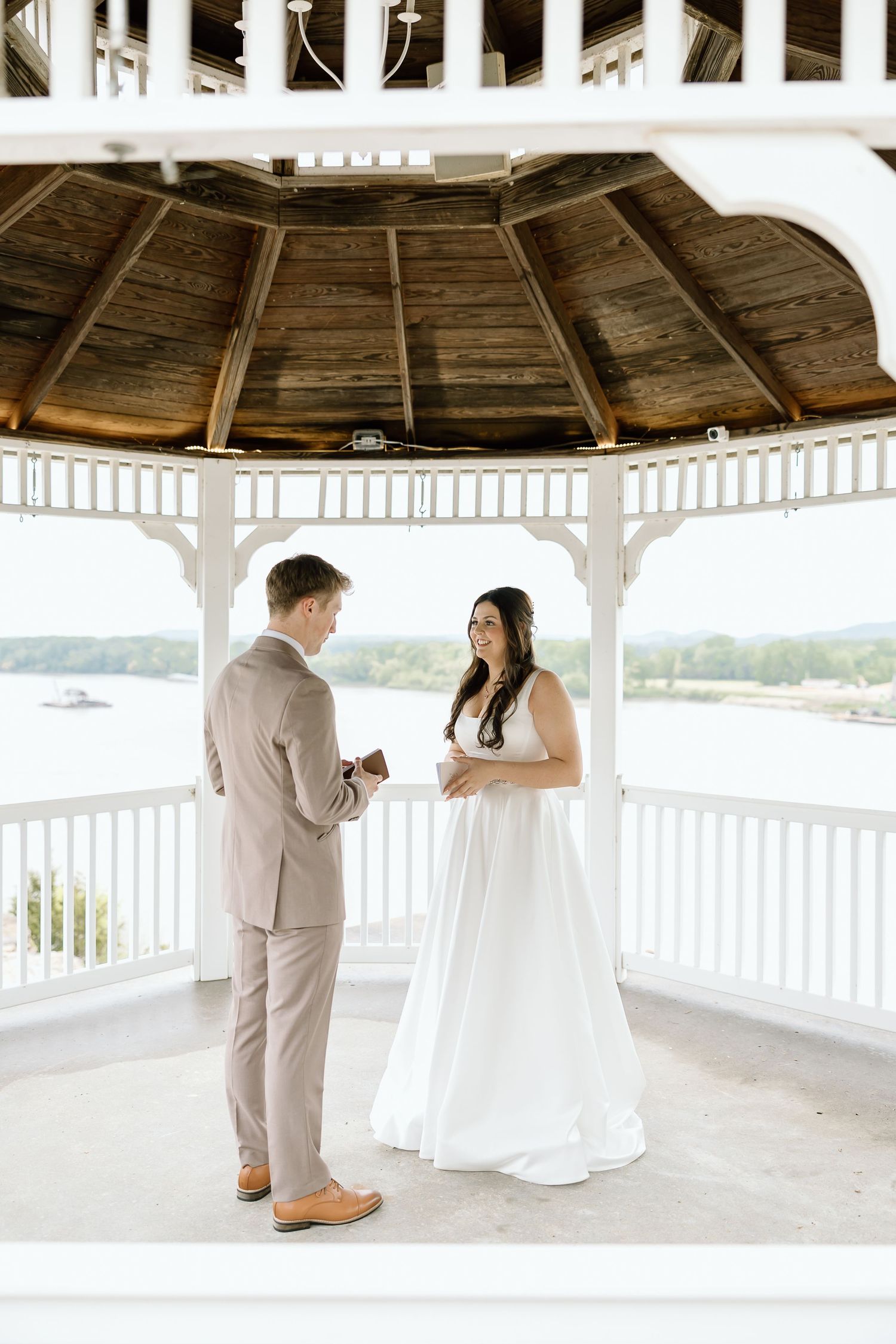 A couple shares a romantic moment in a white wooden gazebo overlooking a waterfront view.