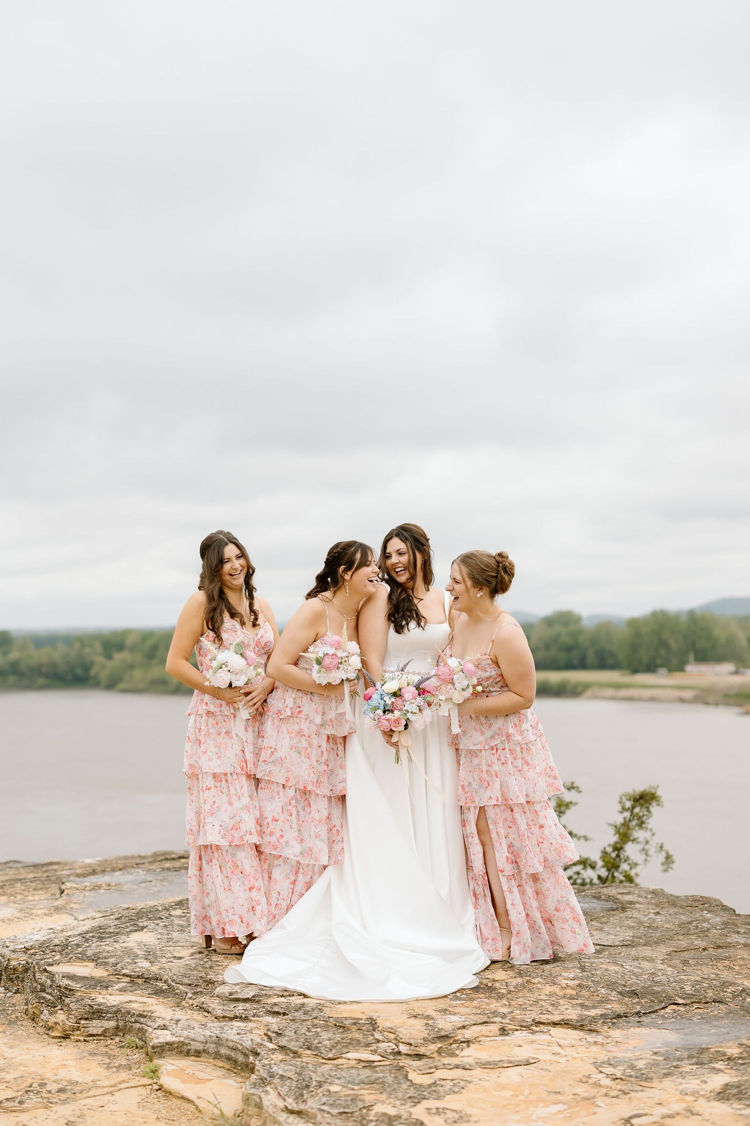 Bridesmaids in floral pink dresses gather with bride on rocky cliff overlooking scenic lake view.