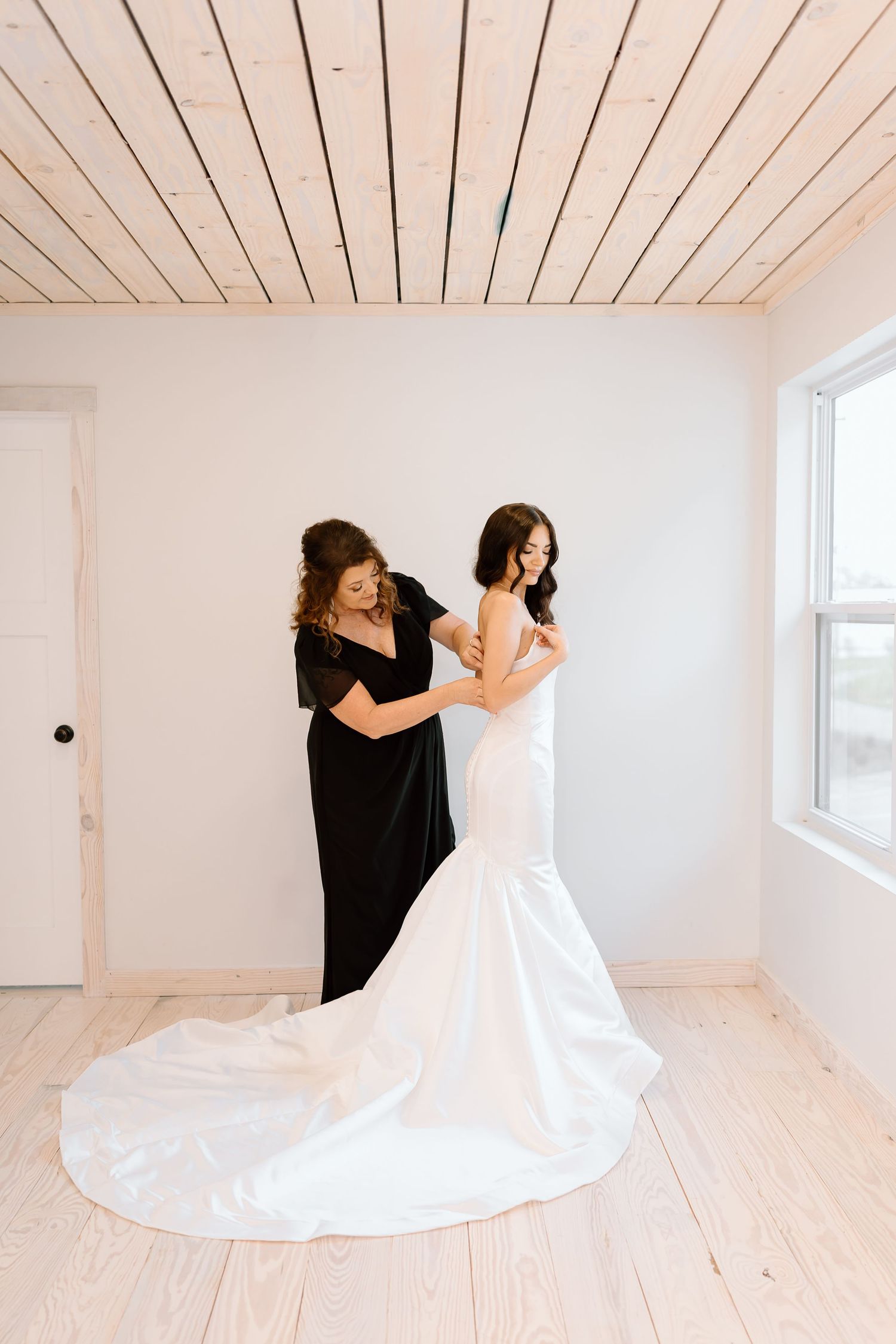 A wedding dress being adjusted in a bright white room with wood plank flooring and natural lighting.