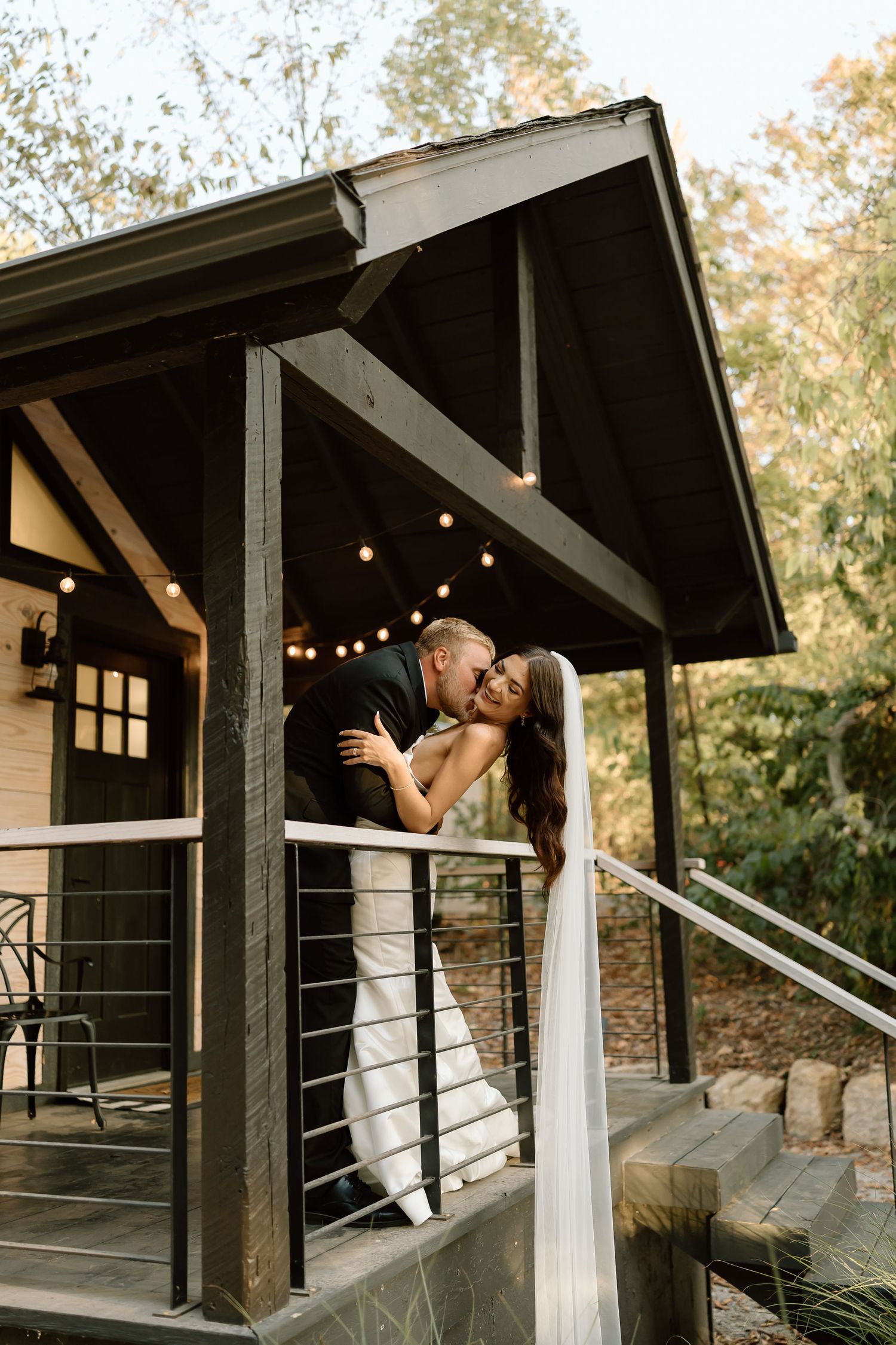 A romantic couple in white attire embraces on a rustic wooden porch with string lights and natural surroundings.