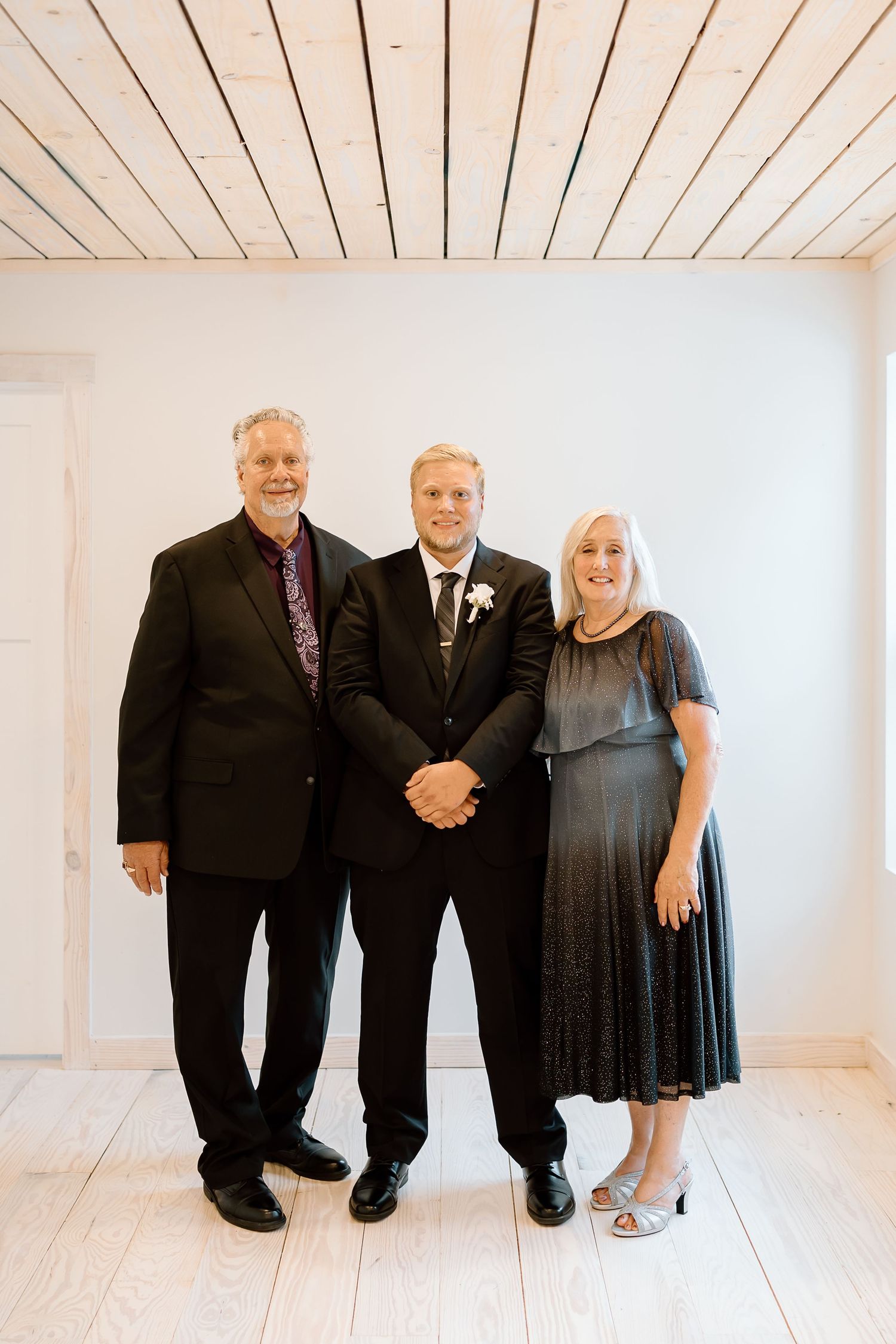 Three people in formal black attire stand together against a white wall in a bright studio setting.