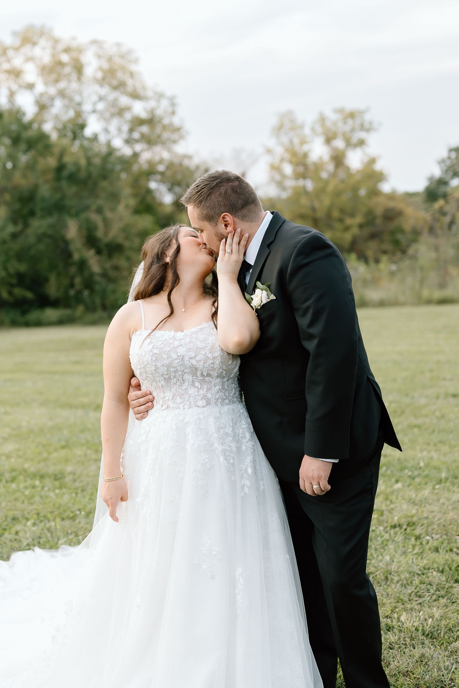 Romantic wedding couple shares tender kisses in a sunlit meadow while wearing formal attire.