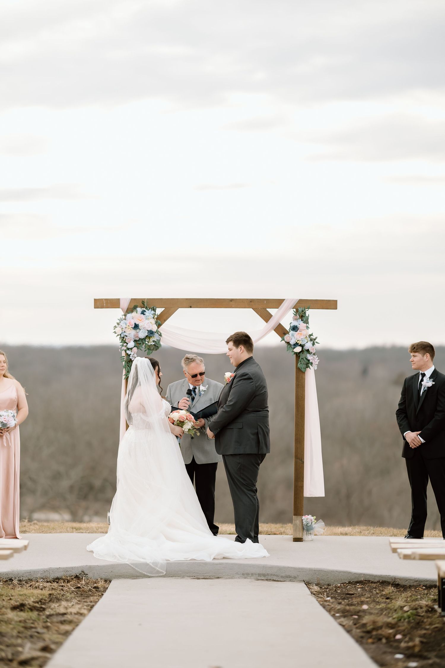 A wedding ceremony takes place at an outdoor venue with a wooden arch and mountain views in the background.