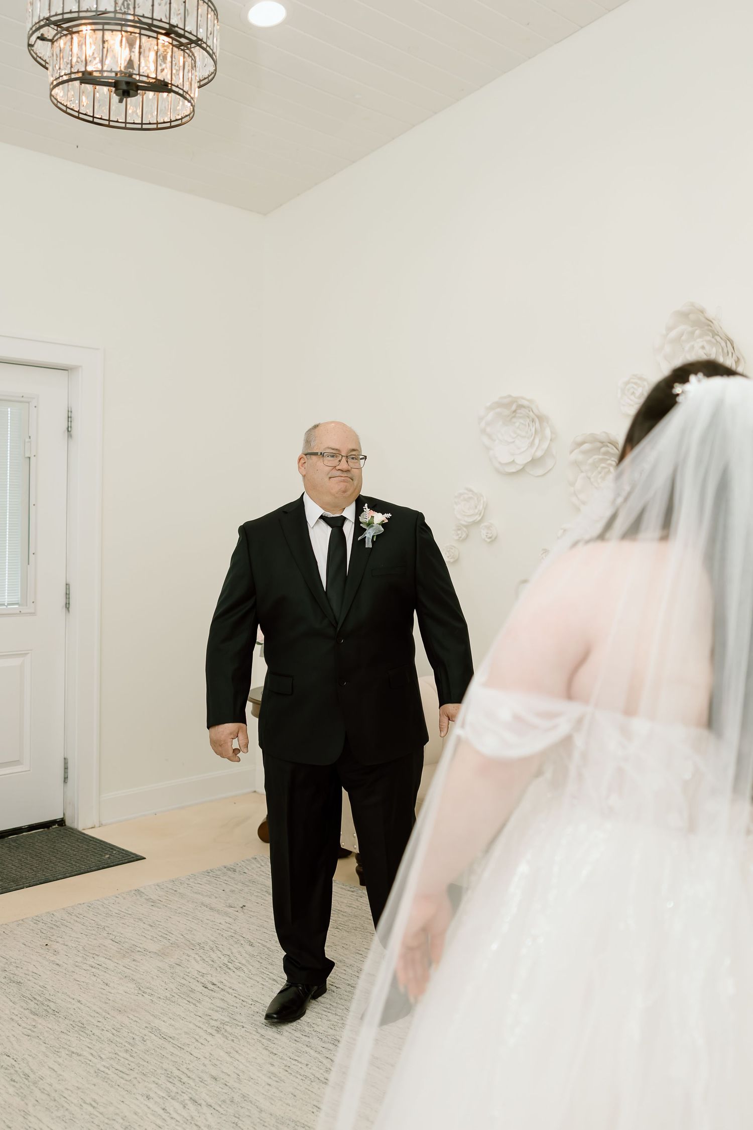 A touching first look moment between a father and bride in a white wedding dress and veil in a bright indoor setting.