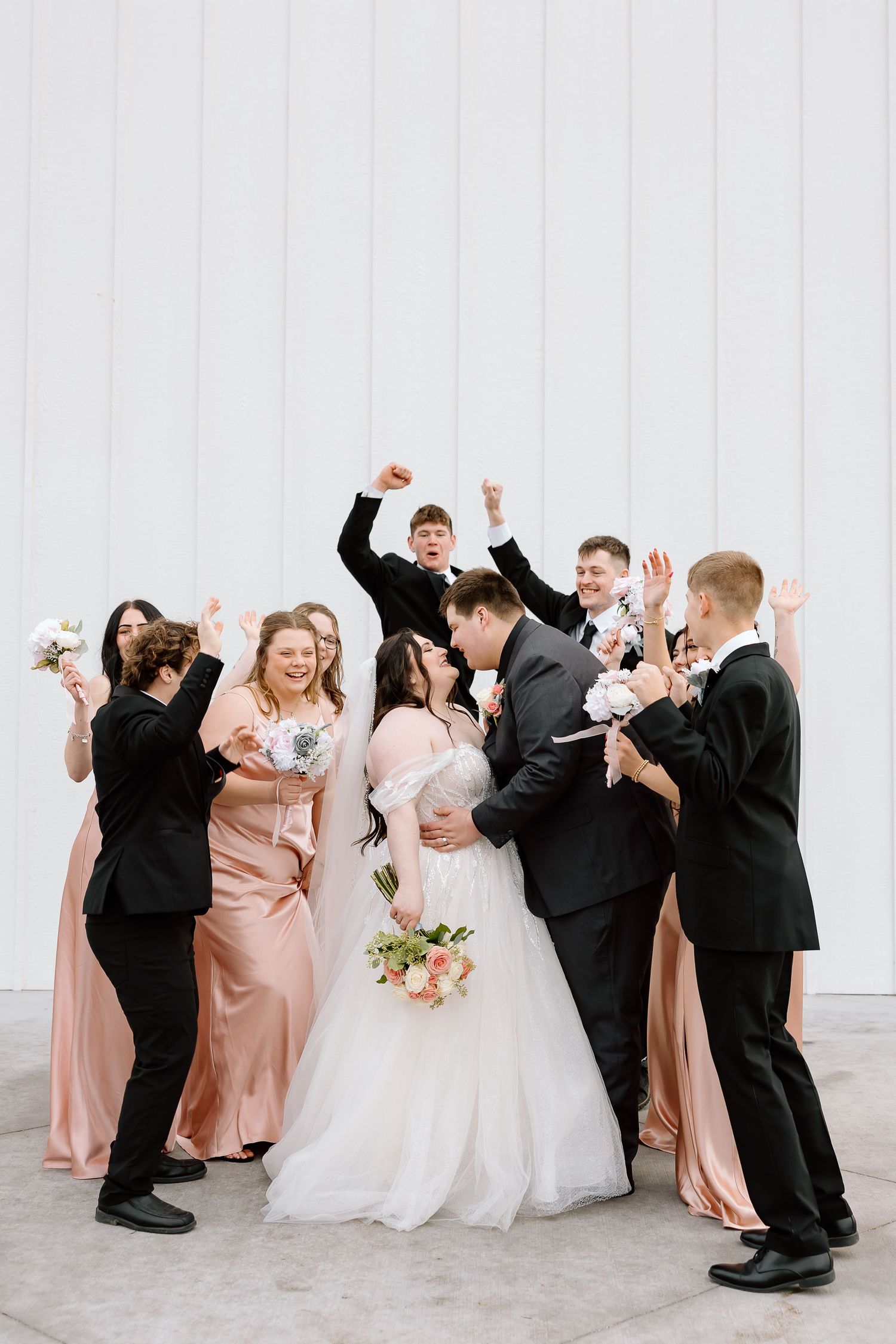 Wedding party celebrates joyfully against white wall with bridesmaids in blush gowns and groomsmen in black suits.