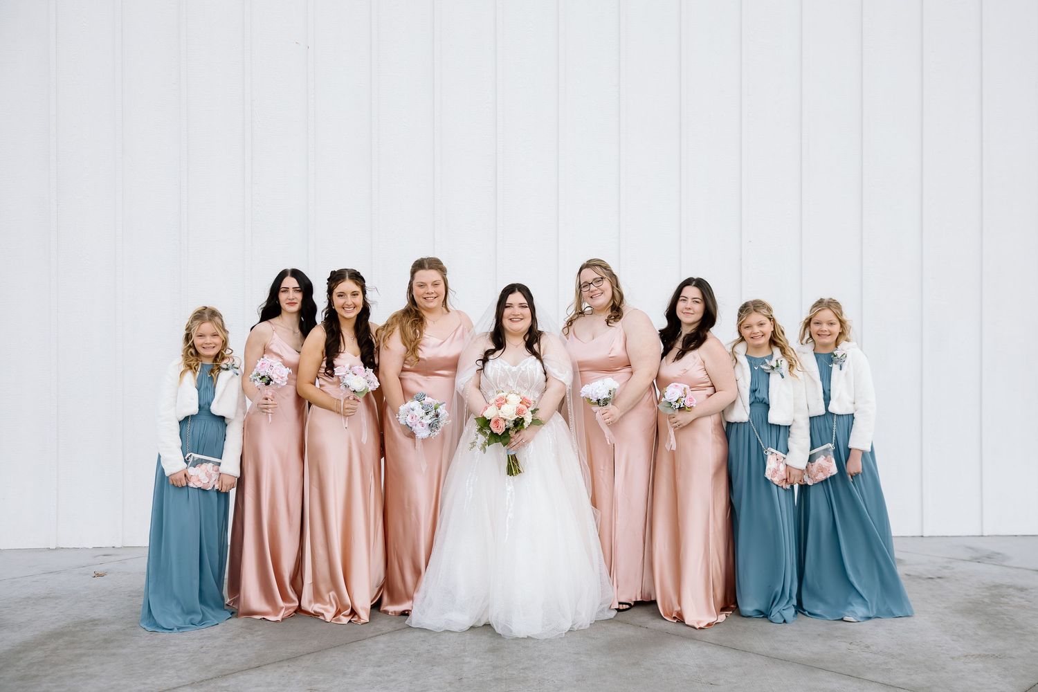 Wedding party members wearing coordinated dusty blue and blush pink dresses pose together in elegant formation.