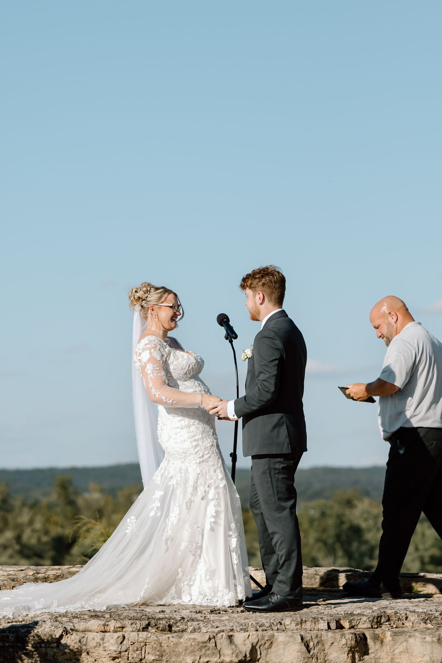 A wedding ceremony takes place on a rocky cliff overlooking a scenic landscape on a clear blue day.