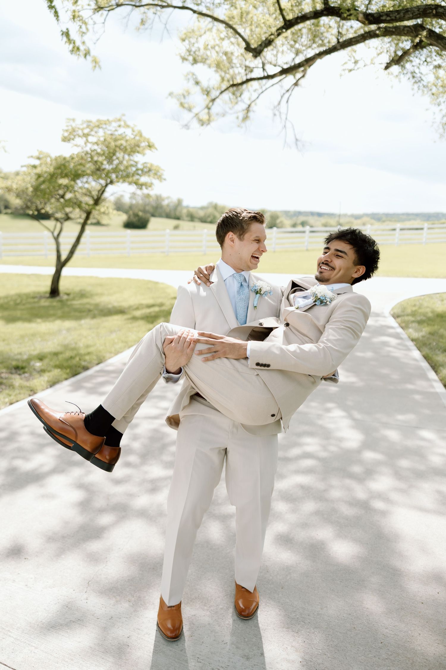 Two grooms in matching beige suits share a joyful moment, one carrying the other on their wedding day outdoors.