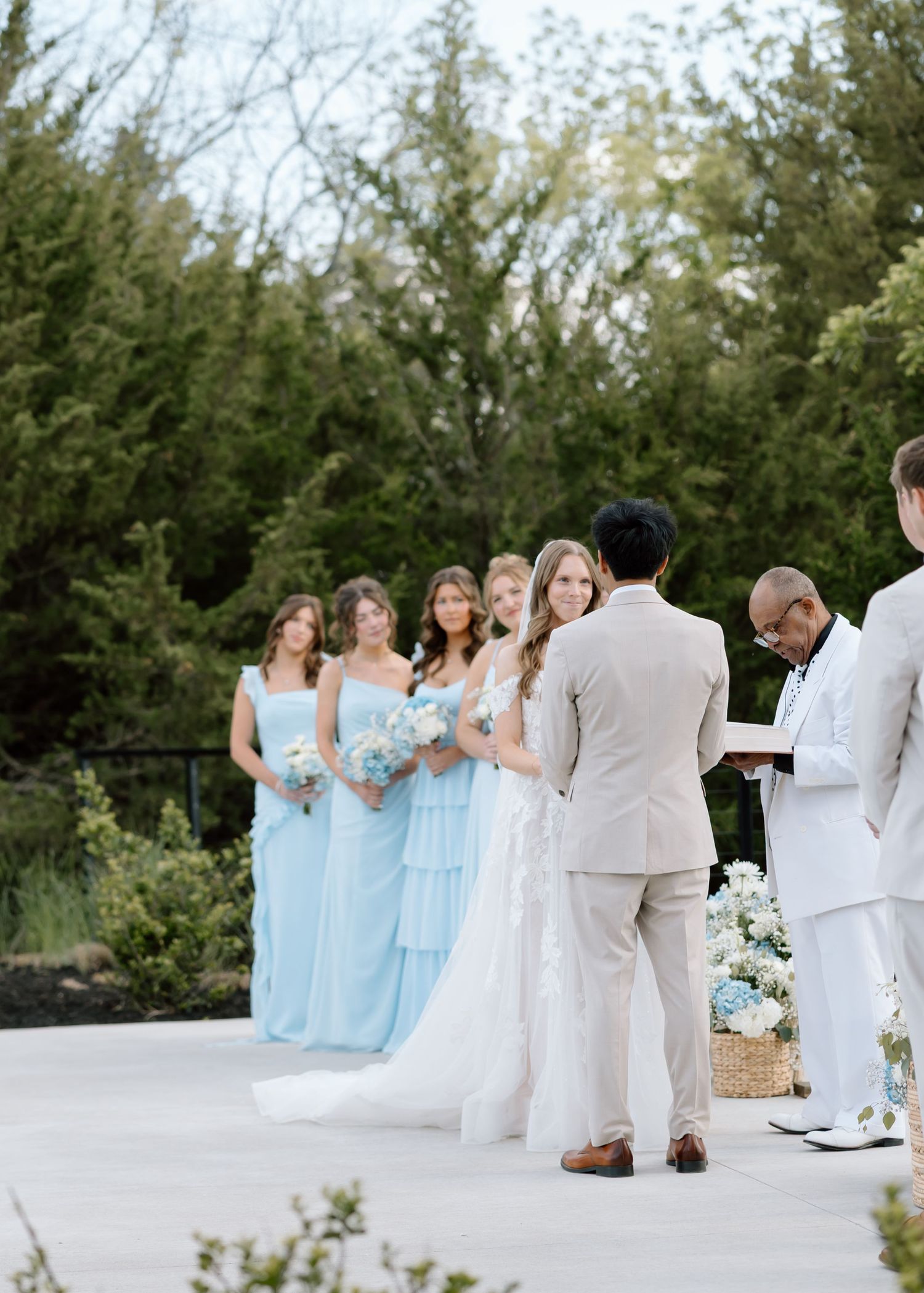 Outdoor wedding ceremony with bride in white gown, bridesmaids in light blue dresses, and groom in tan suit exchanging vows.