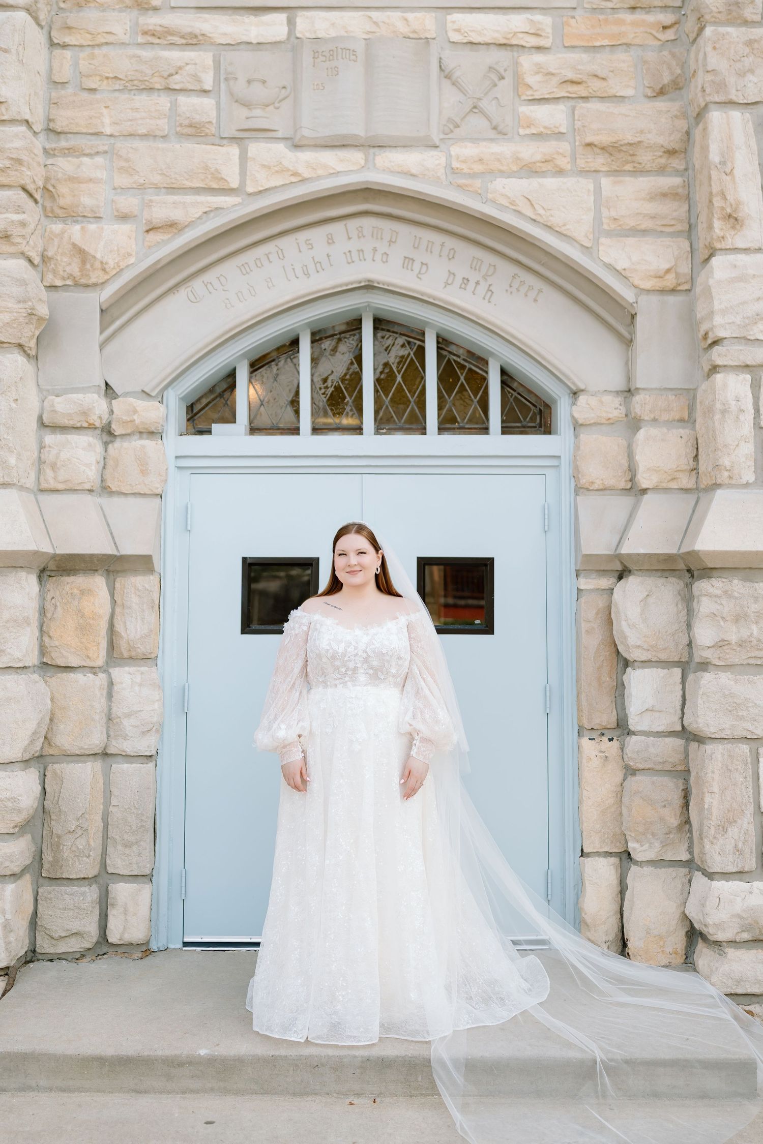 Bride in lace wedding gown with sheer sleeves and veil stands before a stone church with blue doors.