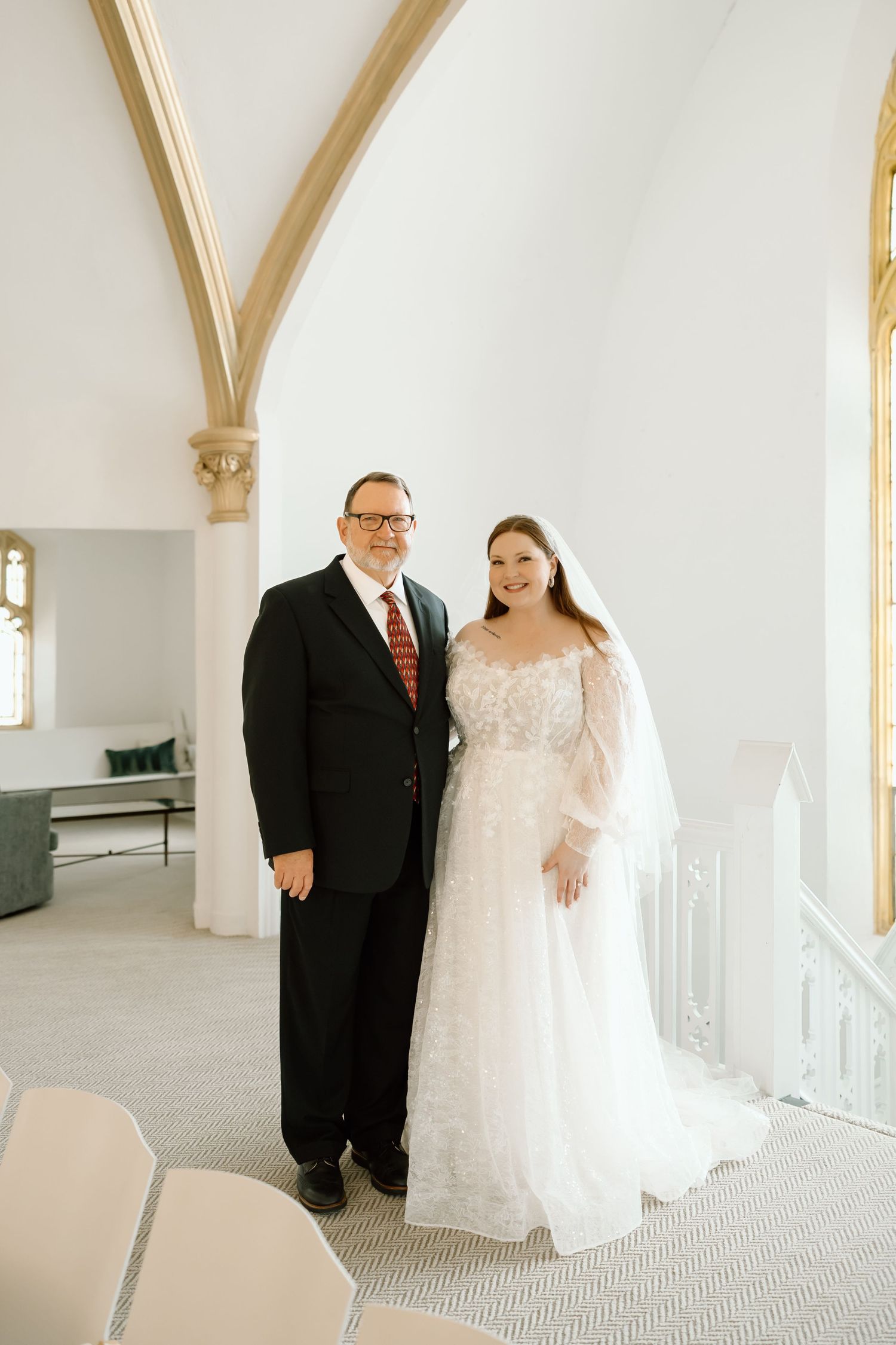 Bride in lace wedding gown with veil stands beside man in black suit inside elegant white chapel.