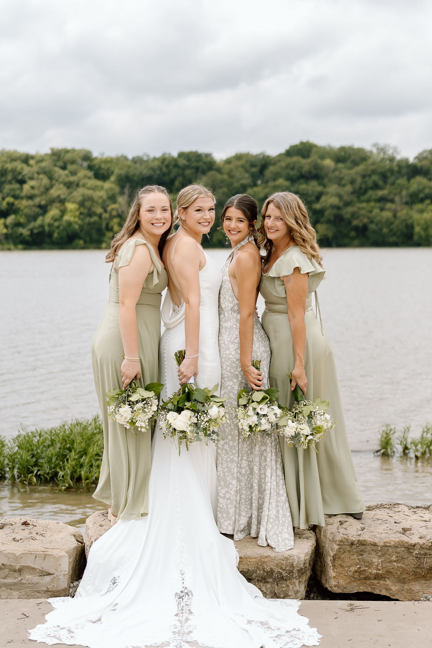 A wedding party poses at a lakeside location with bridesmaids in sage green dresses beside a bride in white.