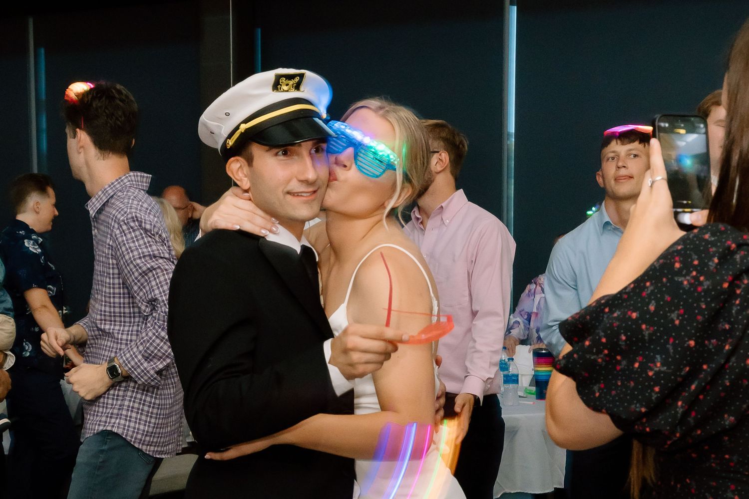 Couples dance together at a dimly lit party venue with people mingling in the background.