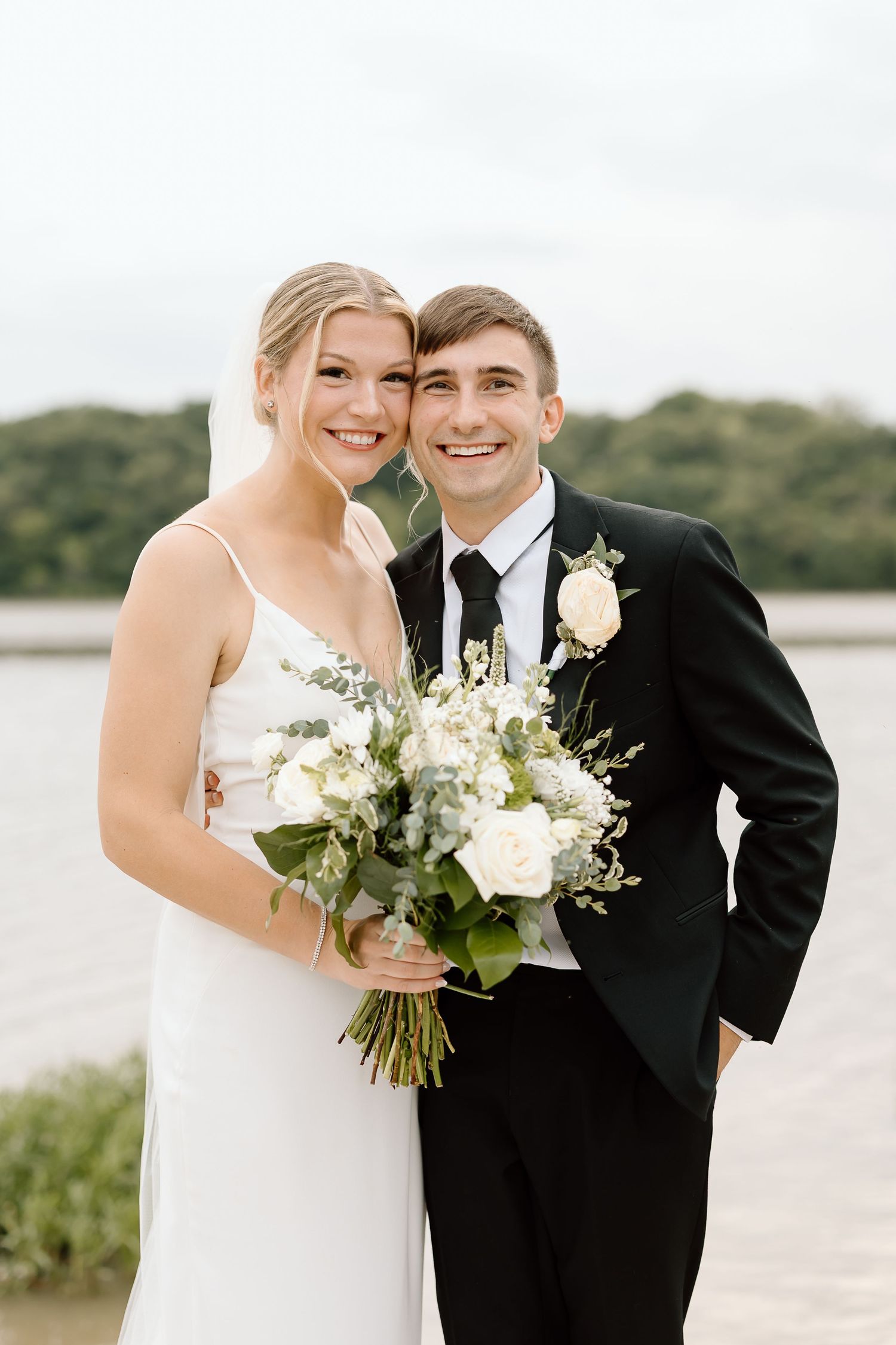 Happy couple shares joyful moments in formal attire by the waterfront with white floral bouquet on their special day.