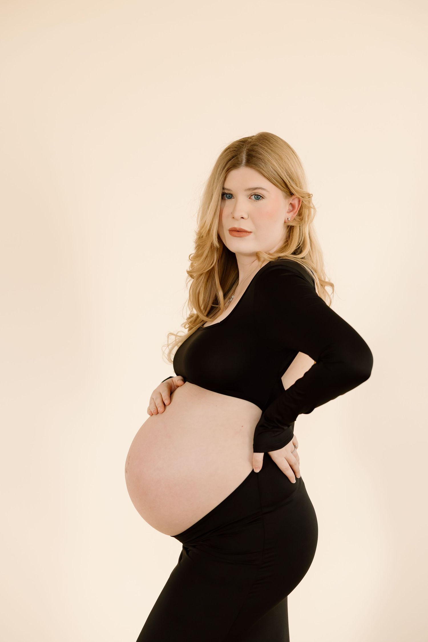 Maternity portrait showing an expectant mother in a black dress posing against a beige background.