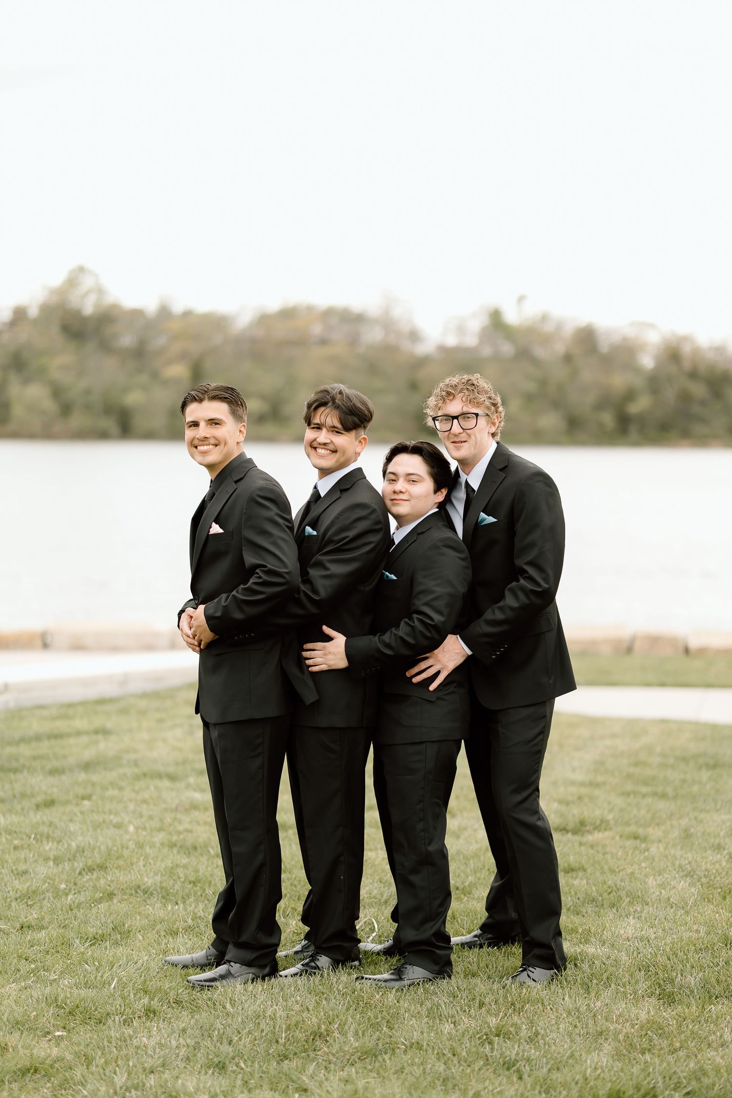 Groomsmen in black suits pose playfully on lakeside lawn.