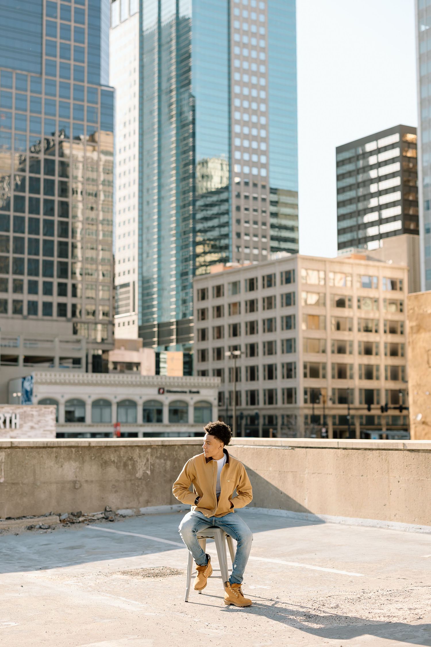 Person in beige jacket sits on stool on urban rooftop with city views.