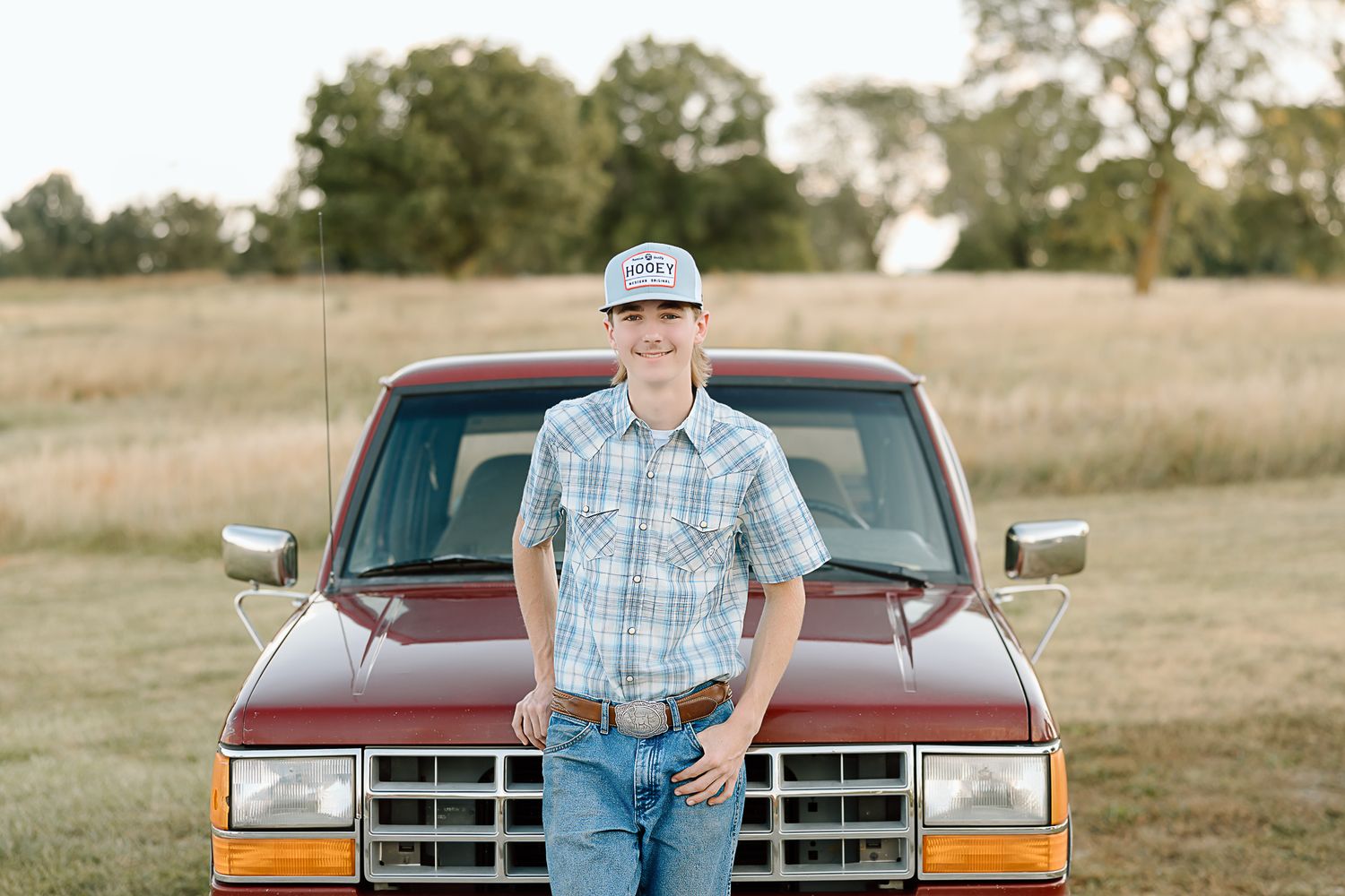 Someone in a plaid shirt and baseball cap poses with a classic red Ford pickup truck in a rural setting.