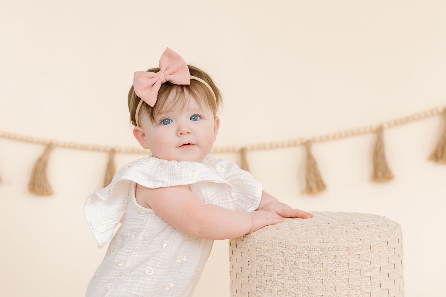 Baby in white dress with pink bow headband stands beside textured stool under tassel garland.
