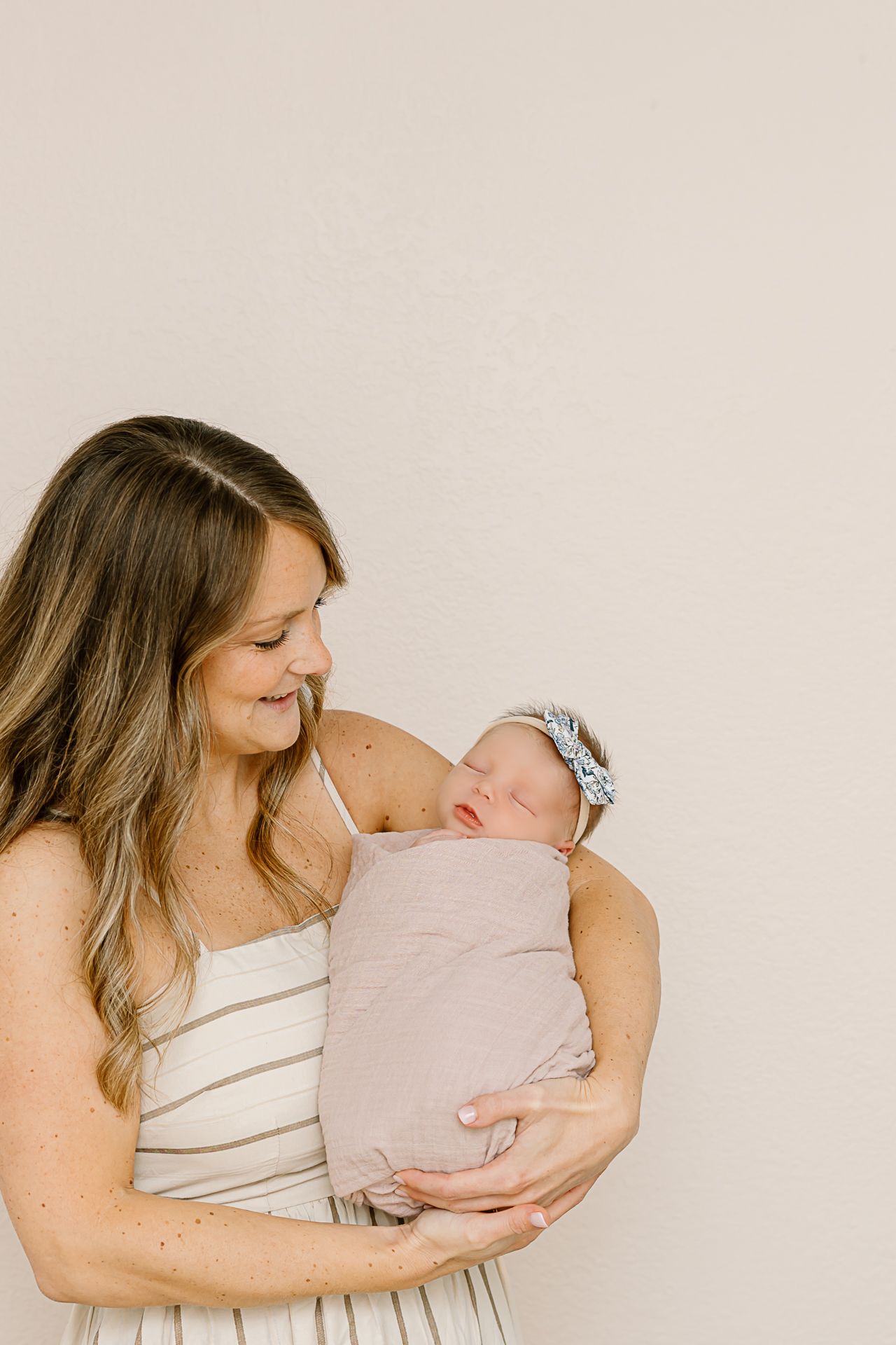 Parent in cream dress holding sleeping newborn against neutral wall.