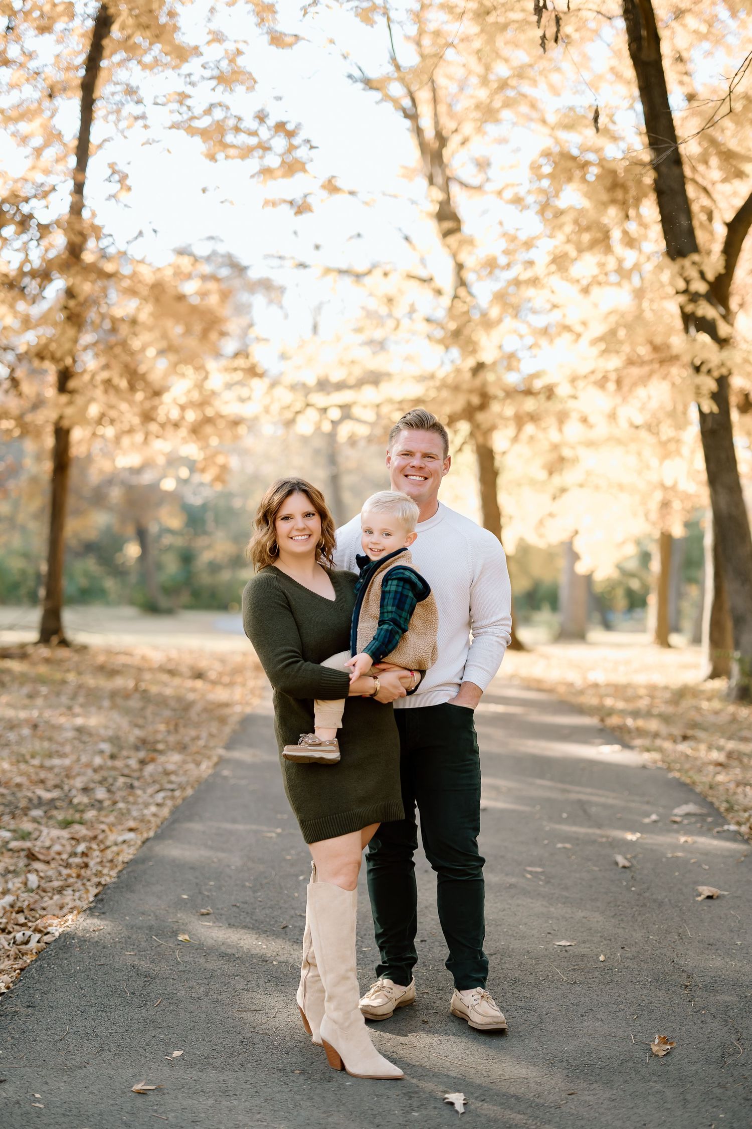 A family of three stands together on a walking path with fall foliage in the background.