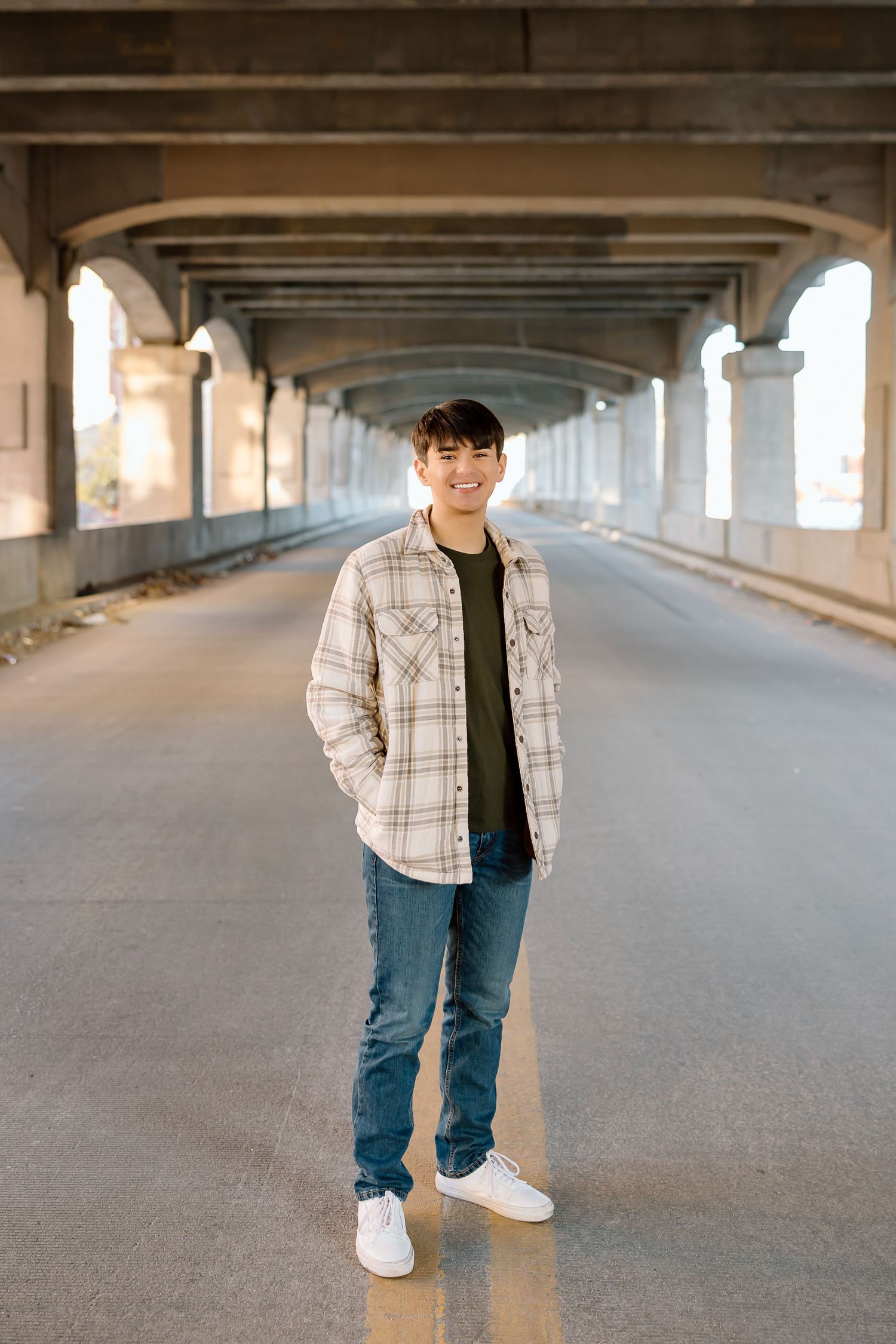 Full length portrait under bridge wearing plaid shirt jacket and jeans.
