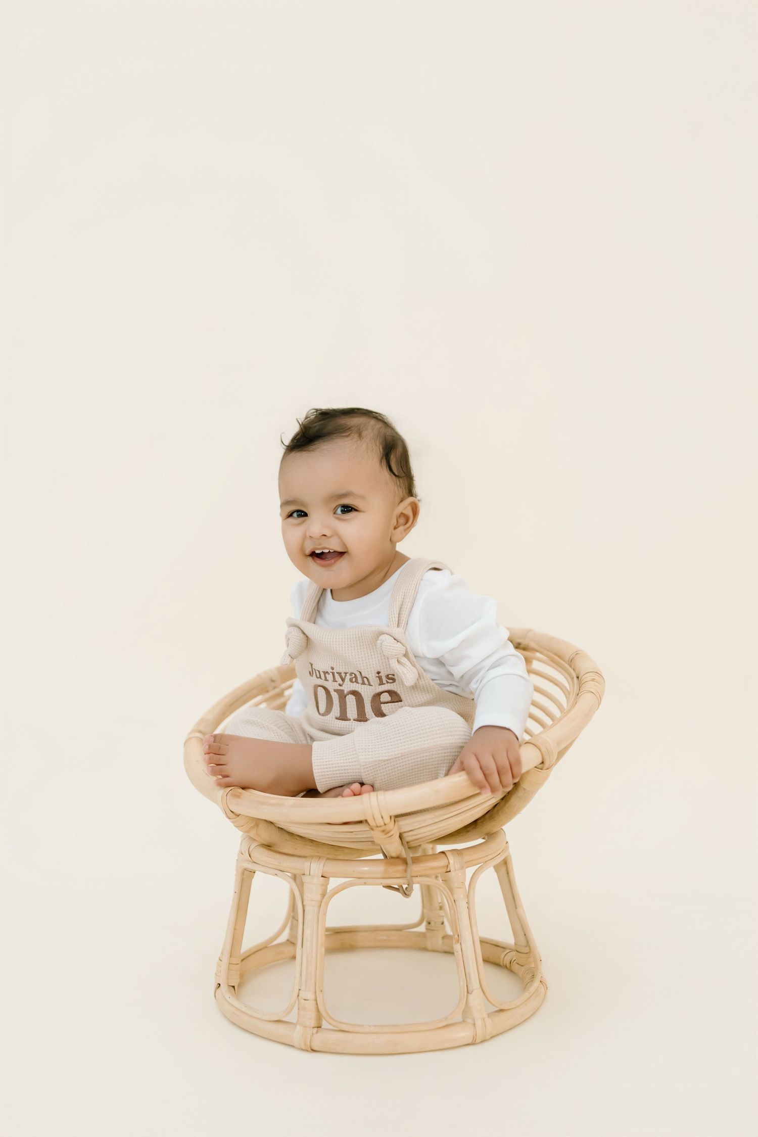 A baby sits in a modern curved wooden rocking chair wearing a white long-sleeve outfit.