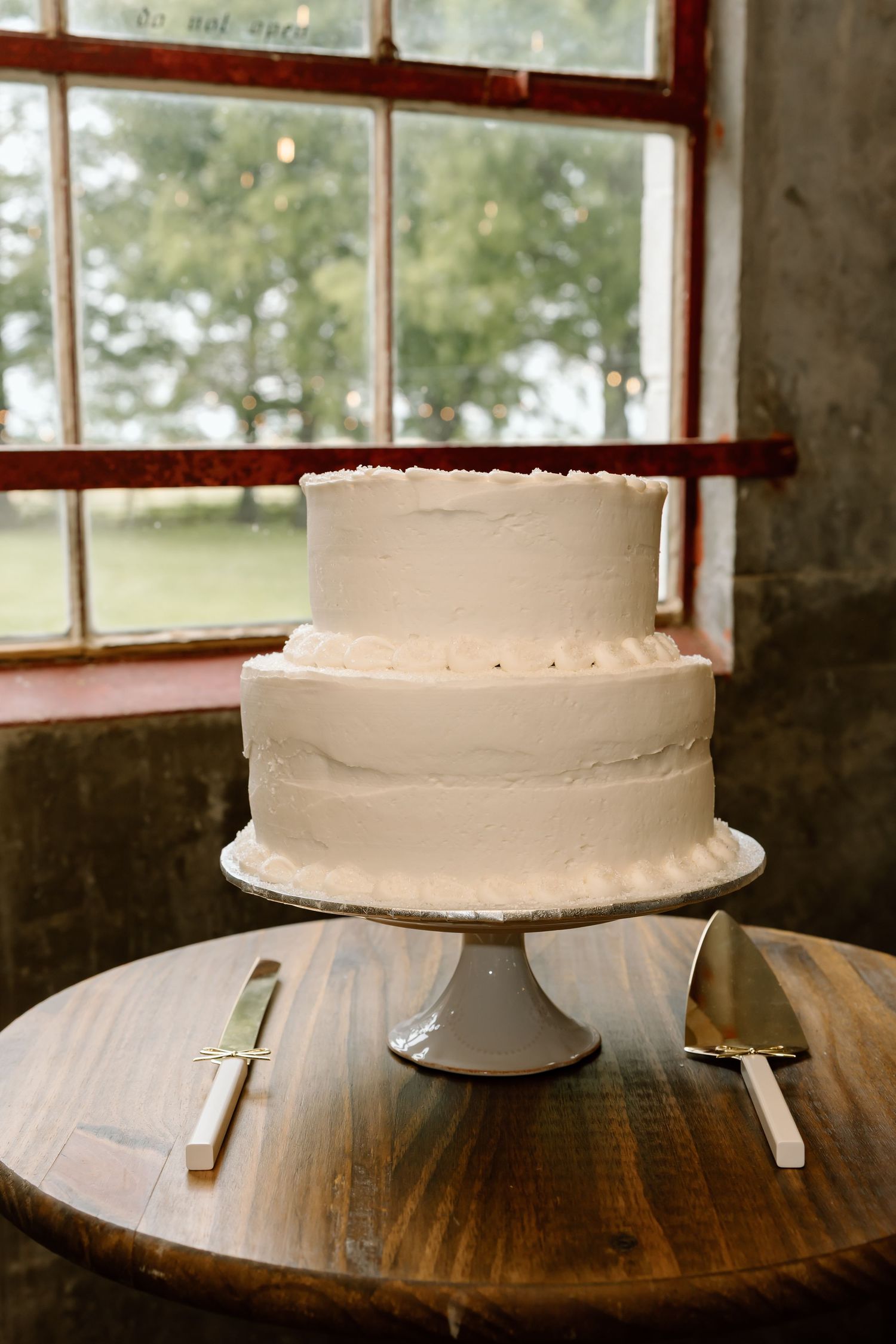 Two-tier white frosted wedding cake on pedestal stand with serving knives on rustic wood table near window.