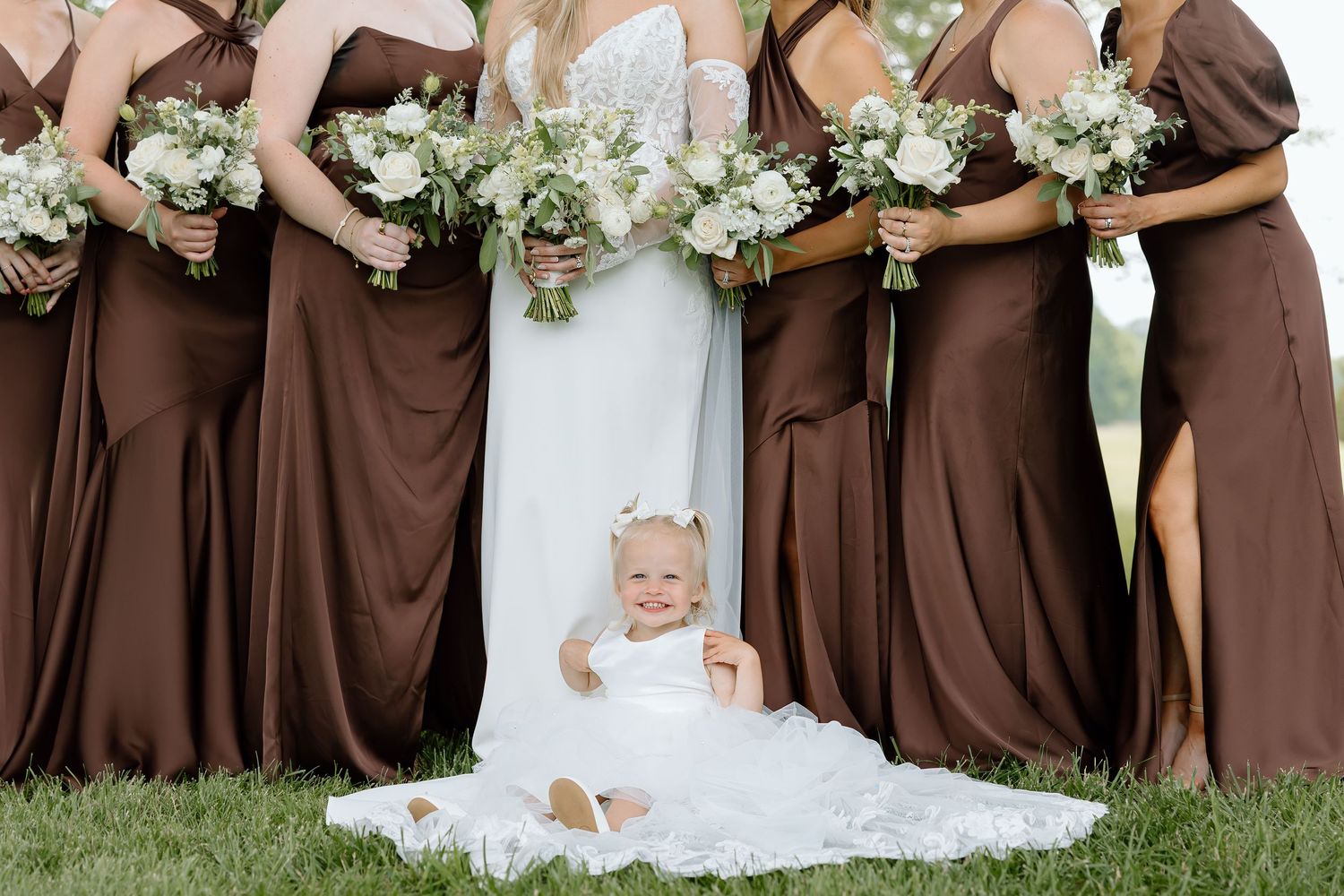 Baby in white dress sits among bridesmaids in brown gowns holding white floral bouquets at a wedding.