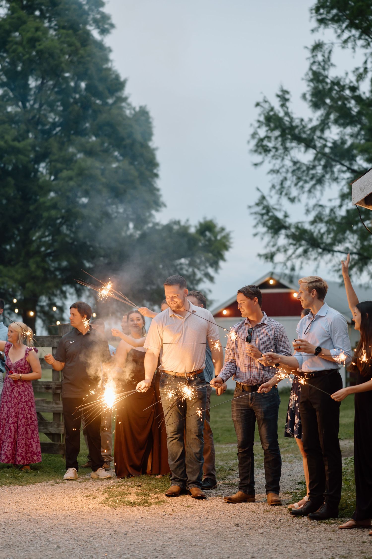 Guests holding sparklers at an outdoor evening wedding reception near a rustic barn surrounded by trees.