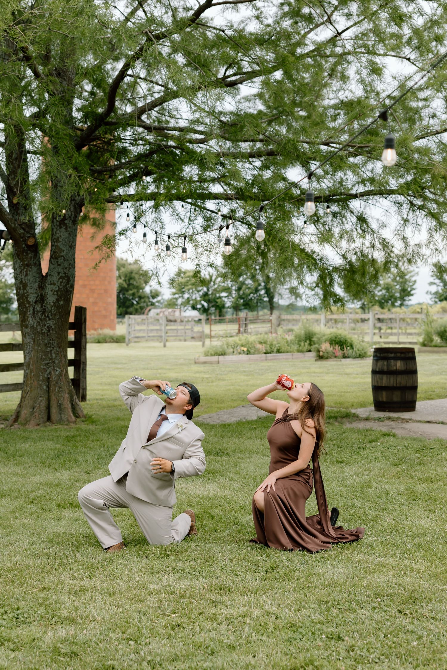 A couple in formal attire kneeling on grass, doing shots outdoors at a rustic venue with string lights and a barrel.