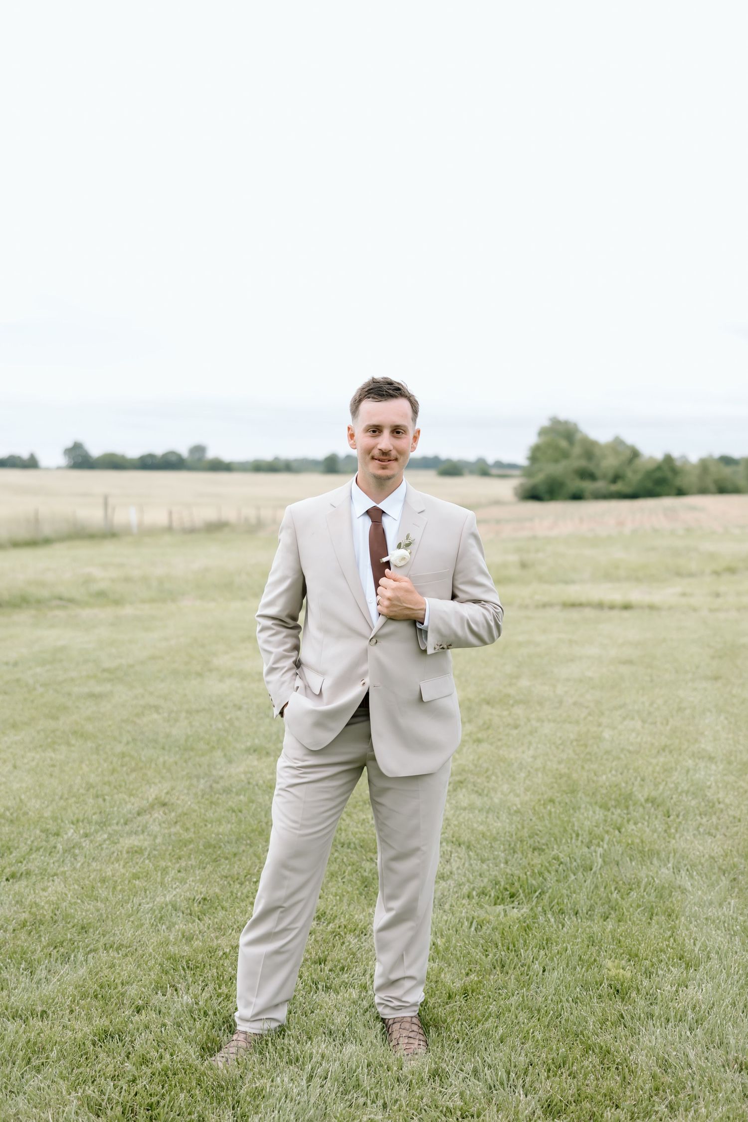 Groom in beige suit with burgundy tie standing in green field smiling at camera on wedding day.