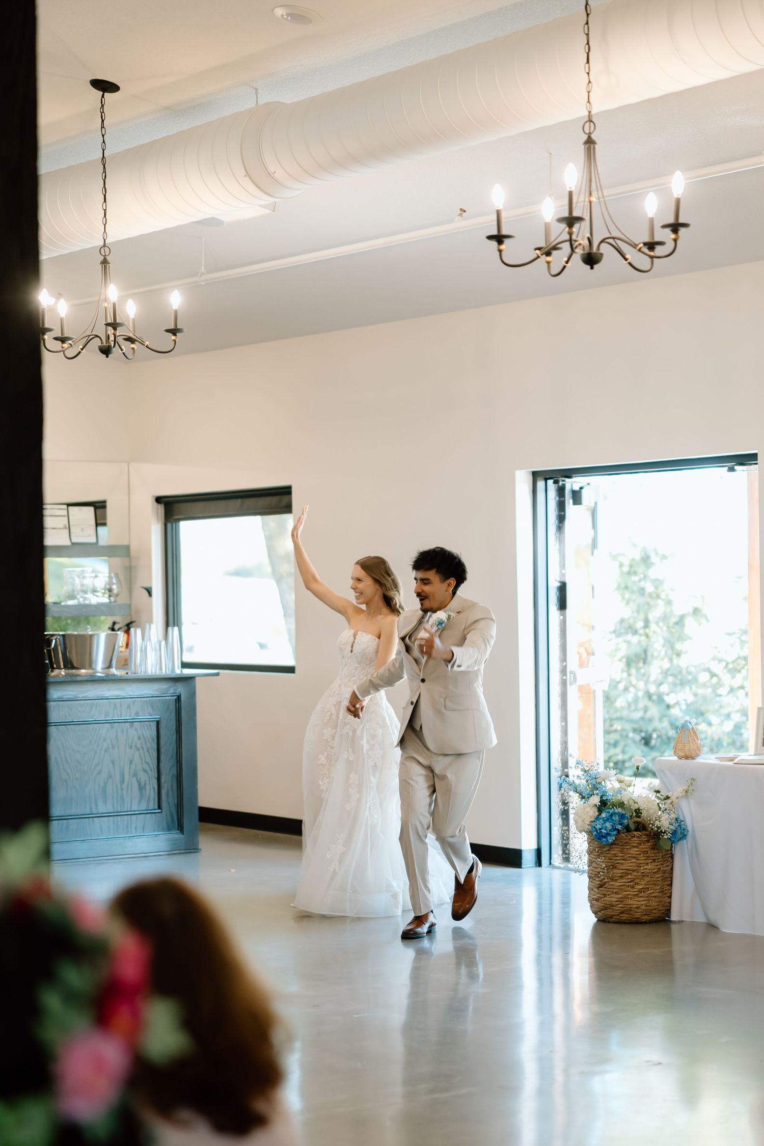 Bride and groom make a joyful grand entrance into their wedding reception hall with chandeliers overhead.