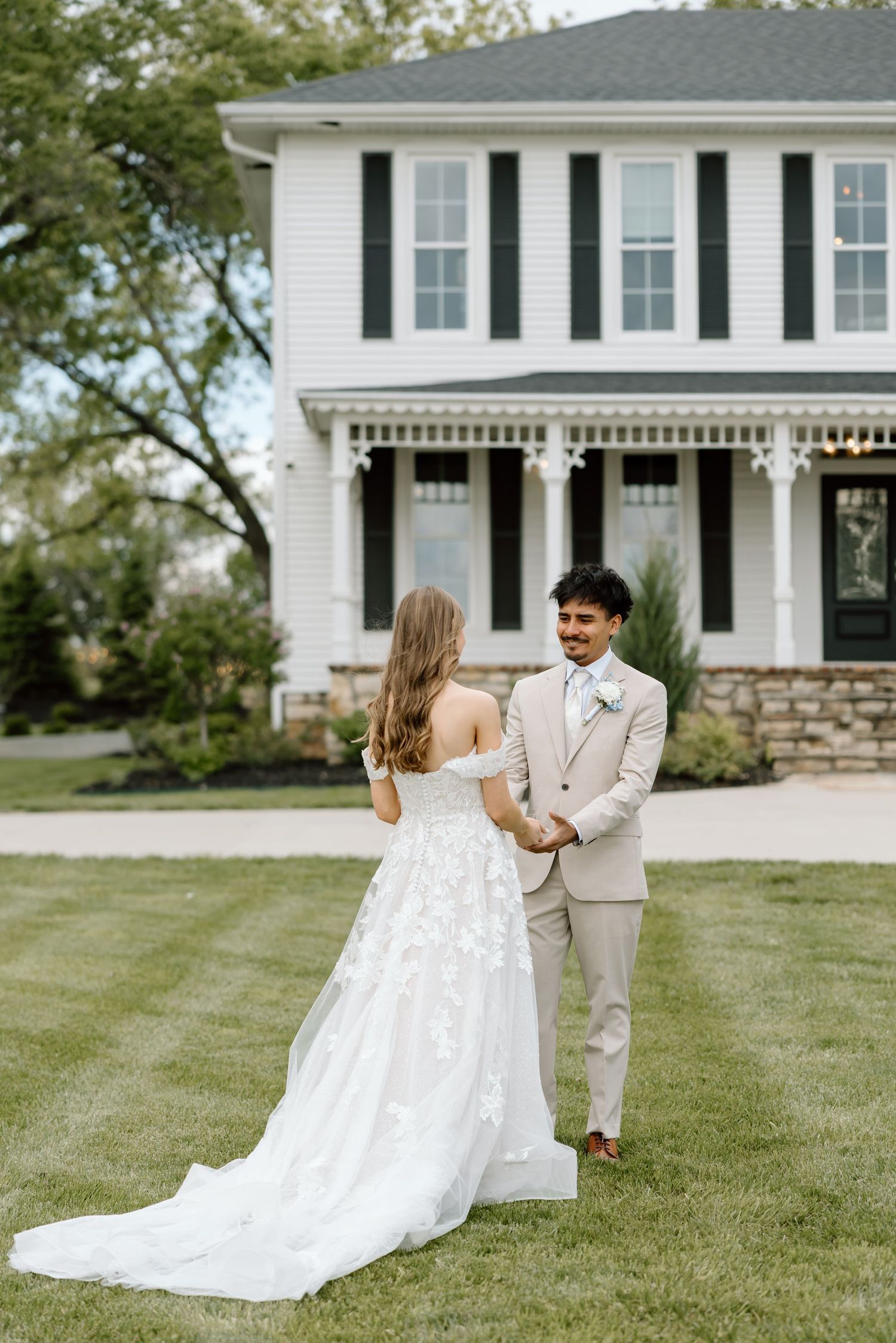 Bride and groom holding hands on green lawn before white farmhouse with black shutters on their wedding day.