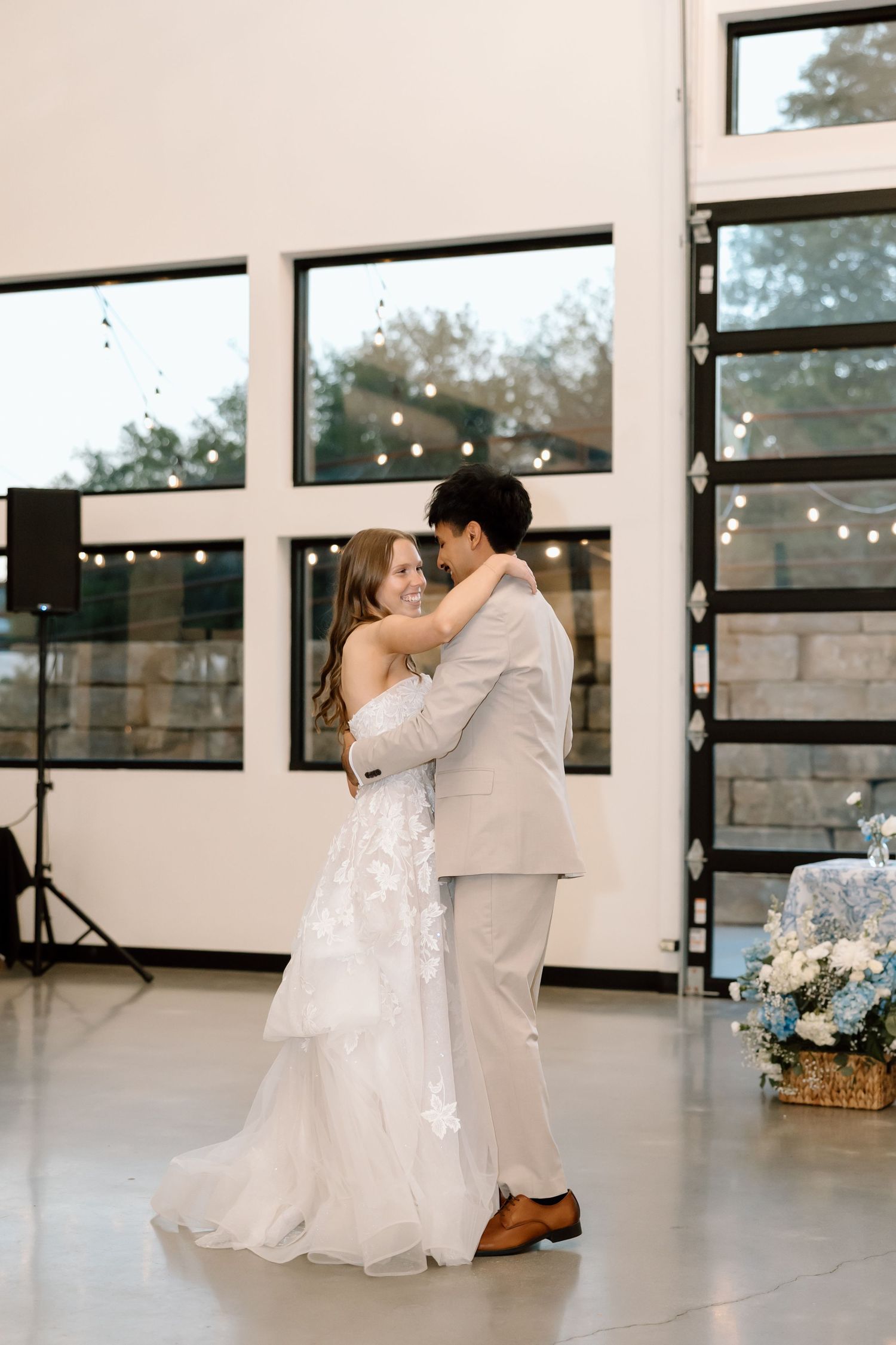 Bride and groom share their first dance in a bright modern wedding venue with string lights.