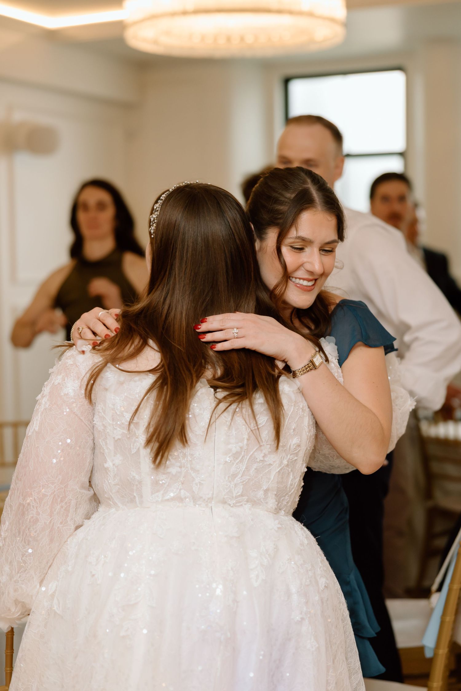 A bride in a white lace gown receives a warm hug from a smiling guest at a wedding reception.
