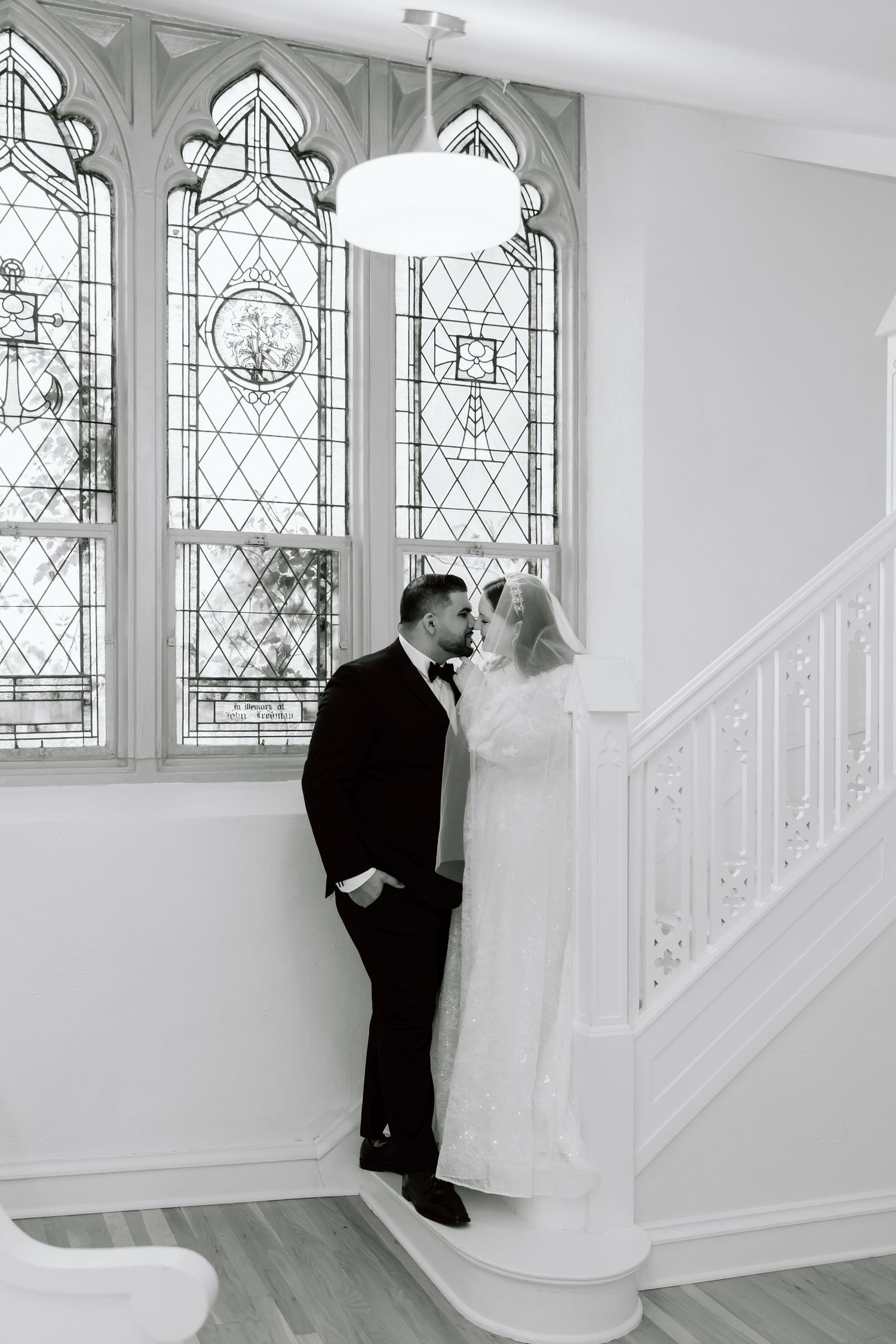 Black and white photo of a bride and groom sharing a romantic moment by stained glass windows near a staircase.